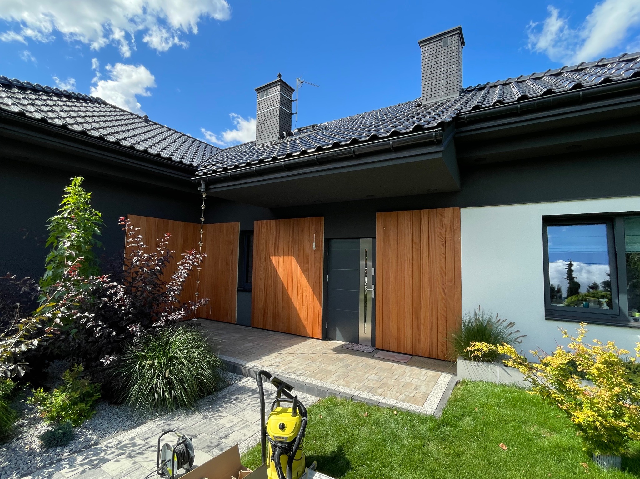 Nowoczesny dom z drewnianą elewacją i kostką brukową. Widoczne rośliny, trawnik i sprzęt czyszczący. Błękitne niebo z chmurami w tle. Słoneczny dzień.