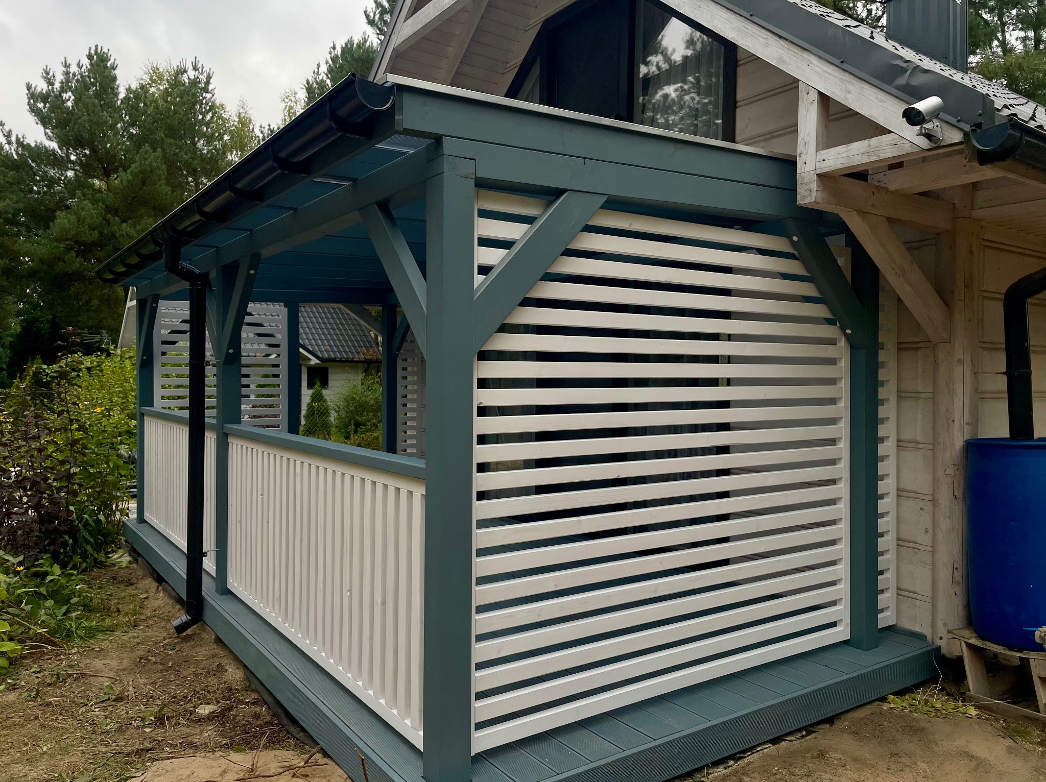 Zadaszona konstrukcja z białymi, poziomymi panelami i elementami w kolorze morskim. Widoczne drewniane słupy i fragment dachu budynku.
