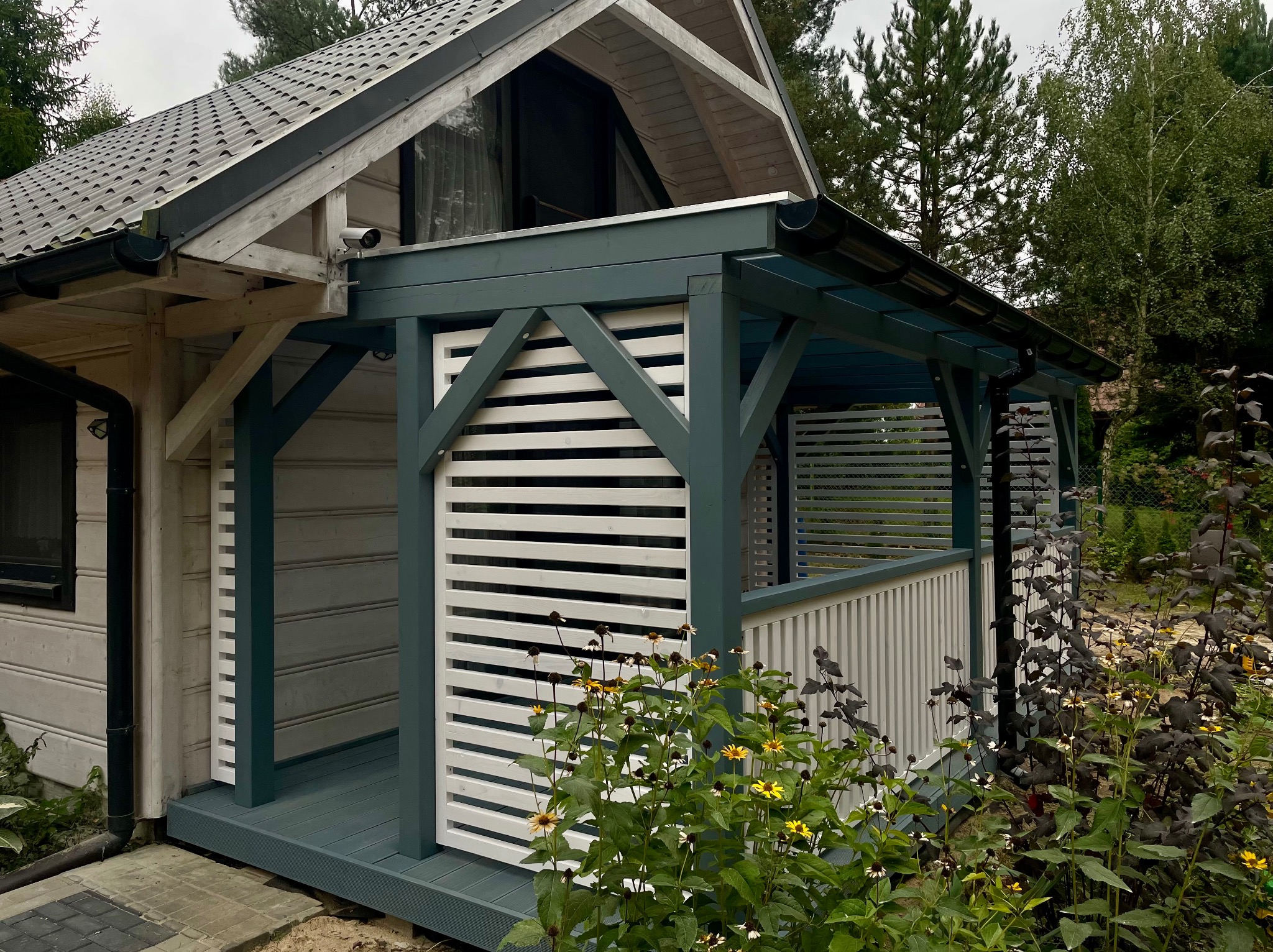 Zadaszona konstrukcja z drewnianymi, białymi lamelami, pomalowana na szaro-zielono. Uroku dodają rosnące przed nią kwiaty. Widoczny fragment domu z kamerą.