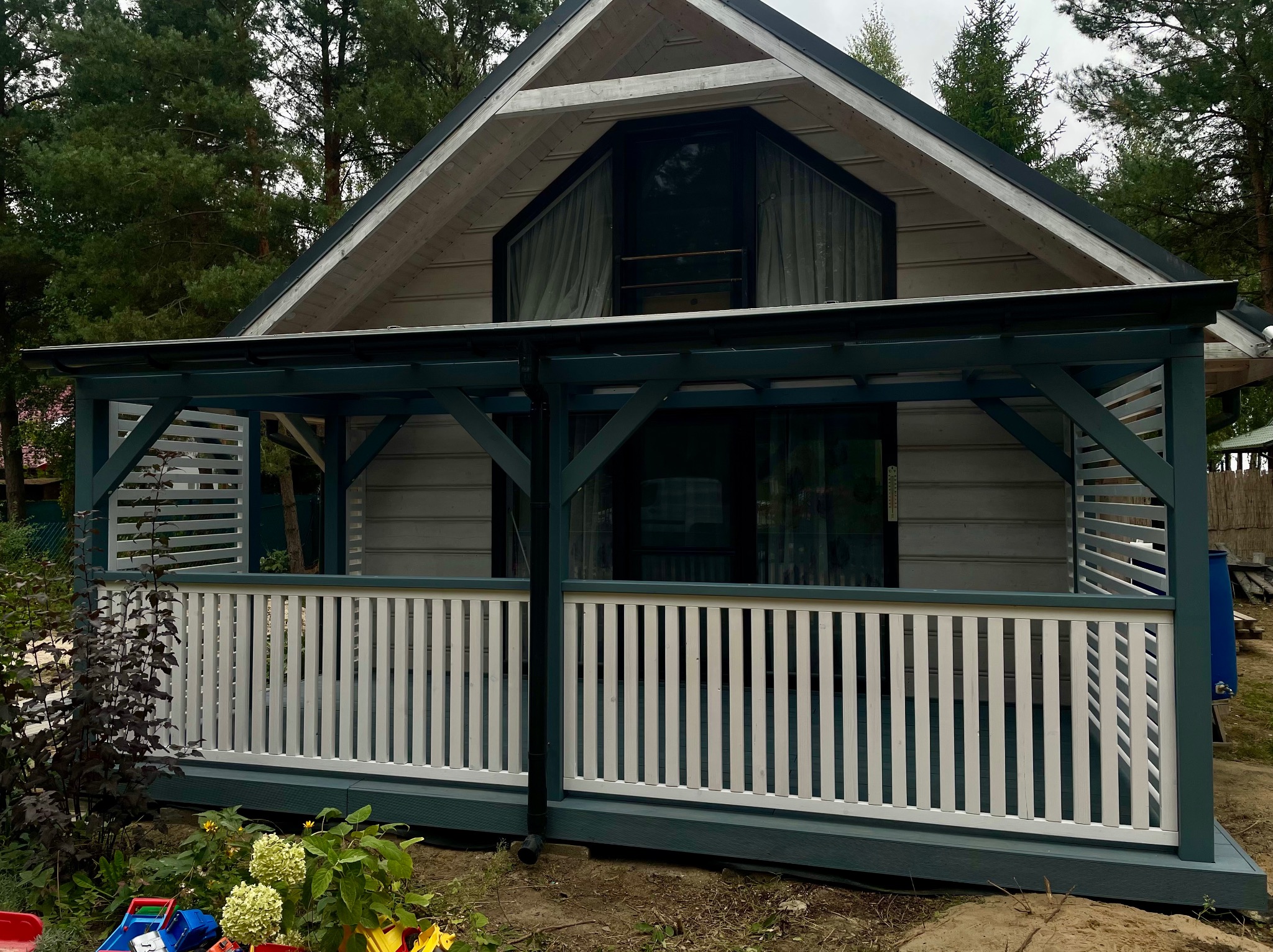 Drewniany domek letniskowy z zadaszonym tarasem i białą balustradą. Elementy konstrukcyjne w kolorze ciemnej zieleni. W tle widoczne drzewa.