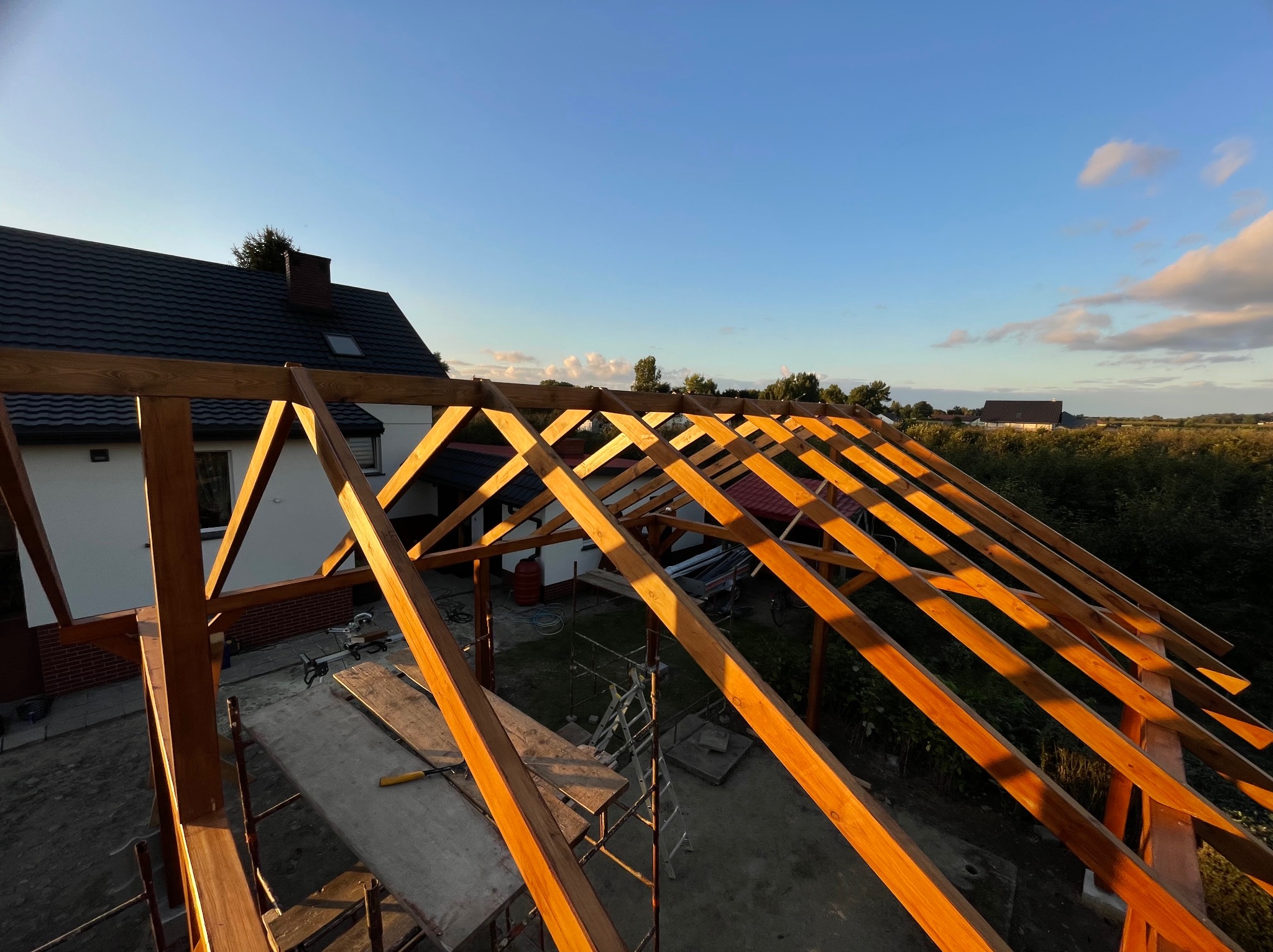 Konstrukcja drewniana z belek w trakcie budowy, widok z góry na dom z ciemnym dachem w tle, rusztowanie i narzędzia pracy widoczne na pierwszym planie.
