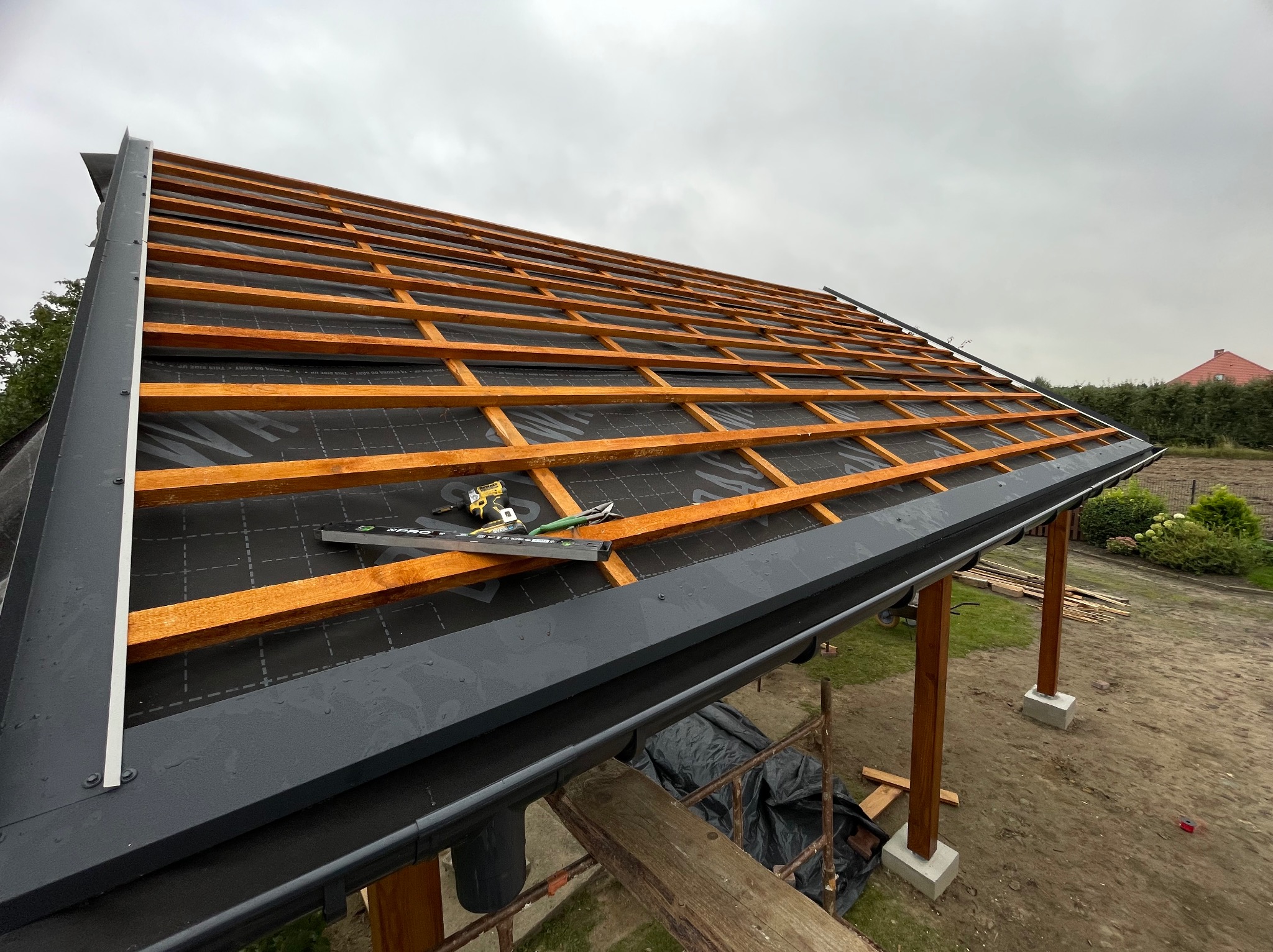 Konstrukcja dachu altany z drewnianymi belkami i czarną membraną, widoczne narzędzia i elementy mocujące, w tle pochmurne niebo. Etap budowy z bliska.