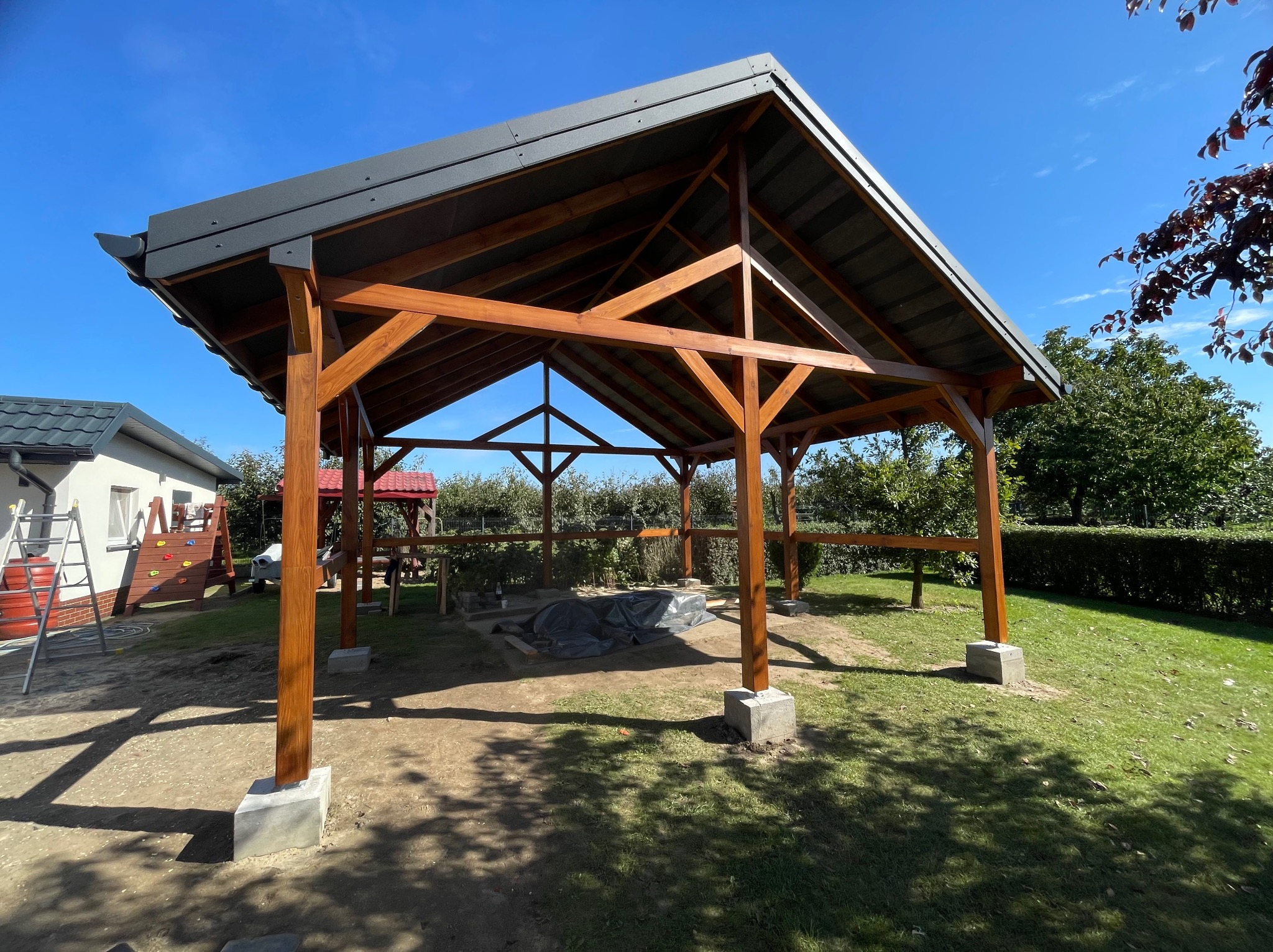 Konstrukcja drewnianej altany z szarym, metalowym dachem na betonowych fundamentach, widok z zewnątrz, w tle zieleń ogrodu i elementy placu zabaw.