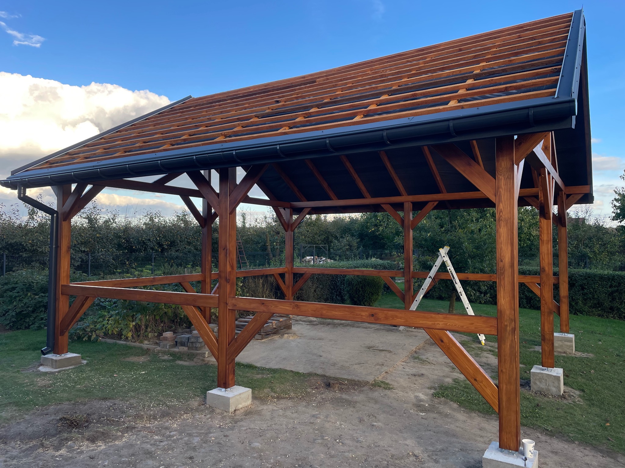 Brązowa drewniana konstrukcja z czarną rynną i częściowo wykonanym dachem na tle zieleni i błękitnego nieba z chmurami, z drabiną w tle.