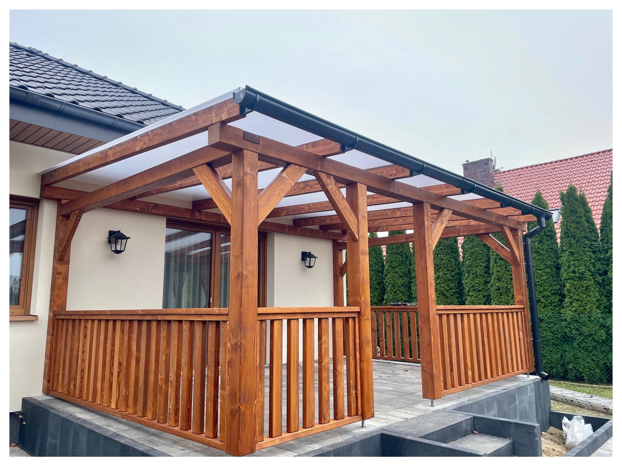 Drewniana pergola z balustradą, zadaszona przezroczystym tworzywem, przylegająca do budynku z oknami i lampami. Widoczne drzewa w tle.