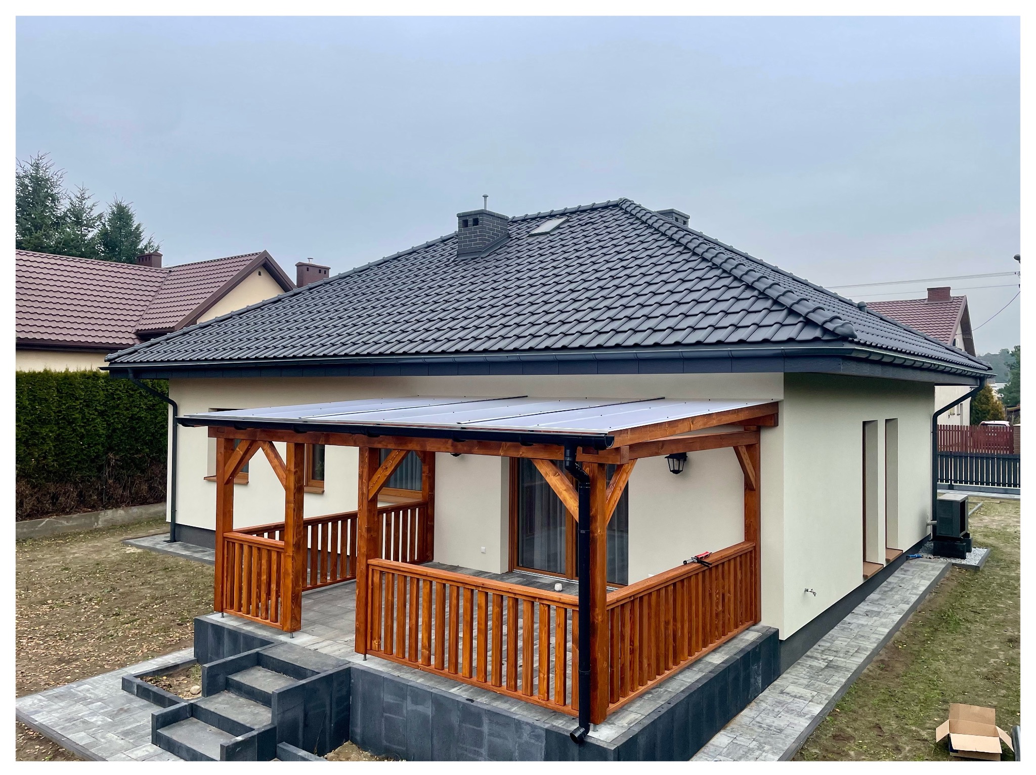 Nowoczesny dom z dobudowaną, zadaszoną konstrukcją z drewnianą balustradą i schodami wejściowymi z kostki brukowej. Dach pokryty ciemną dachówką.