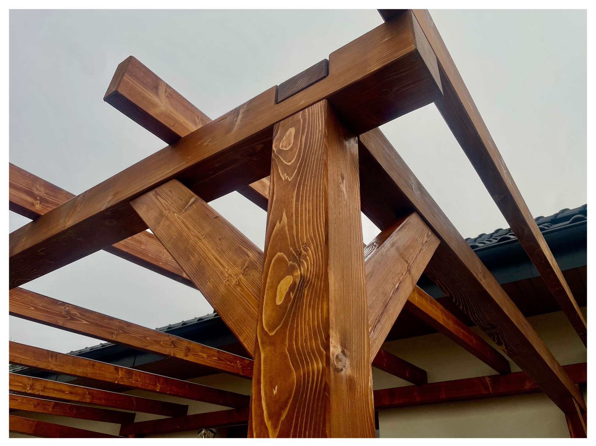 Brunatna, drewniana konstrukcja pergoli z widocznymi detalami łączeń, na tle pochmurnego nieba i fragmentu dachu. Widok z dołu, podkreślający solidność wykonania.