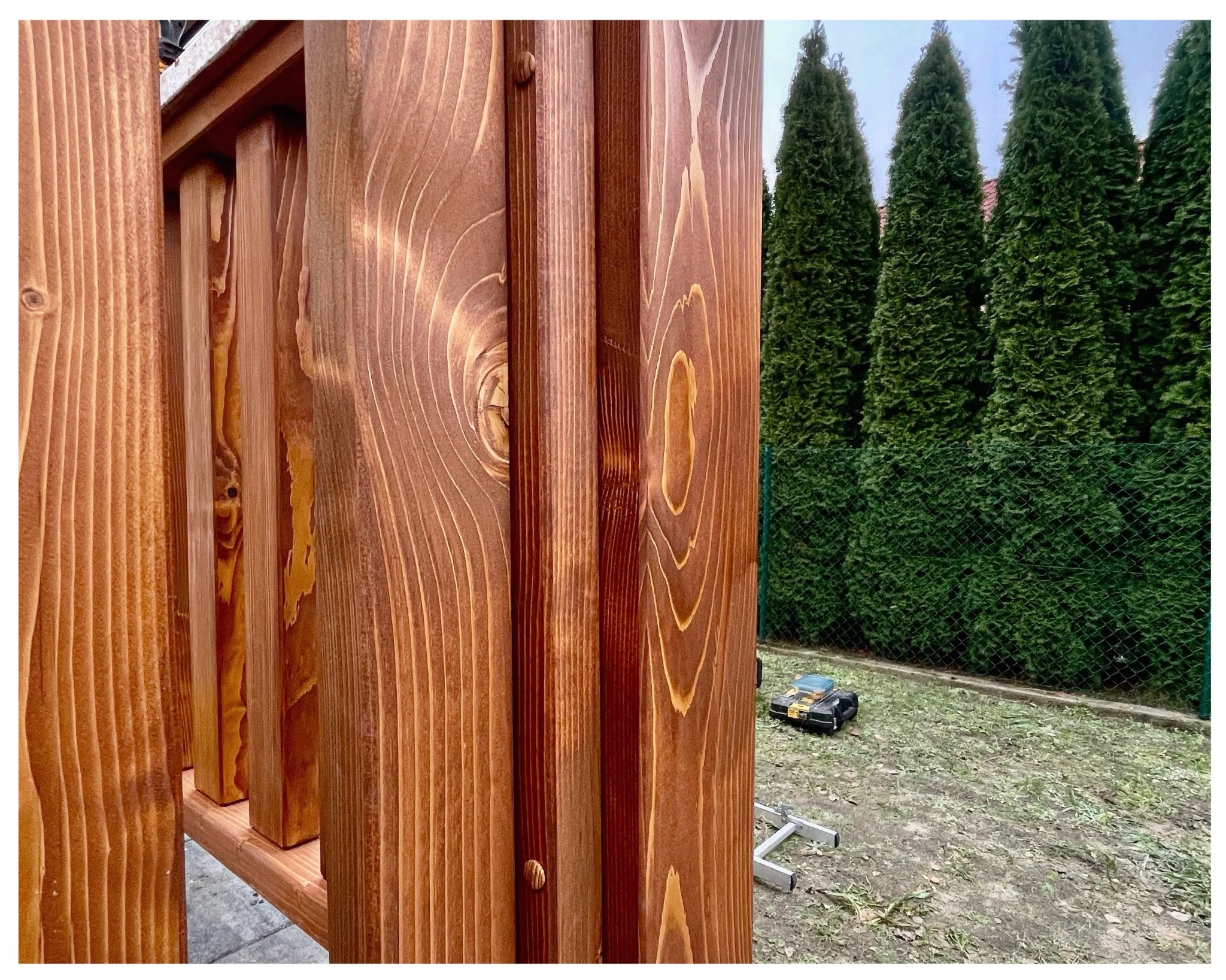 Fragment konstrukcji z brązowego drewna z widocznym usłojeniem, na tle zielonych drzew i trawnika z narzędziami. Detal wykończenia, ciepłe barwy drewna.