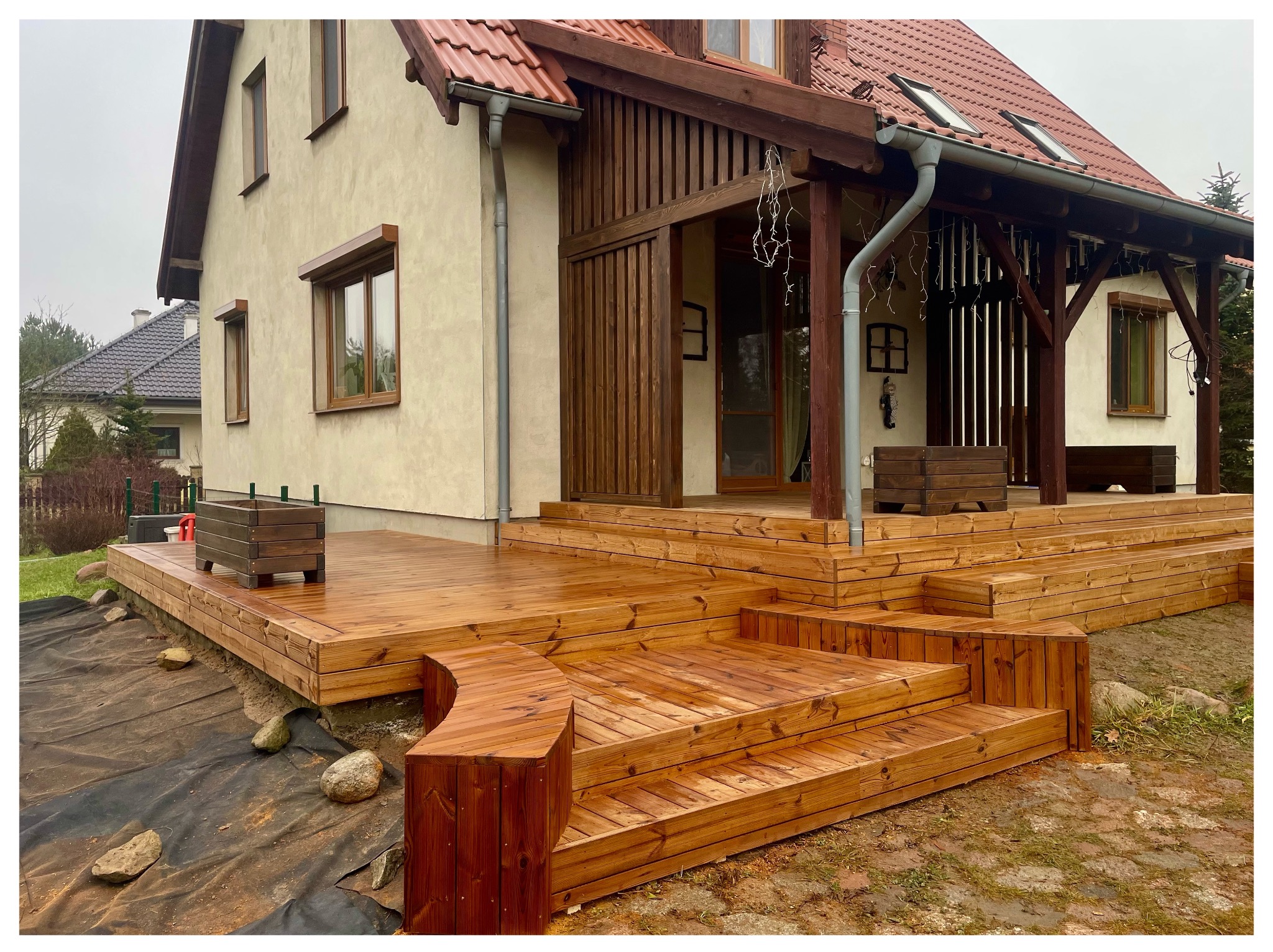 Drewniany taras z schodami i donicami, przylegający do domu z drewnianą werandą. Widoczne elementy wykończeniowe i detale architektoniczne.