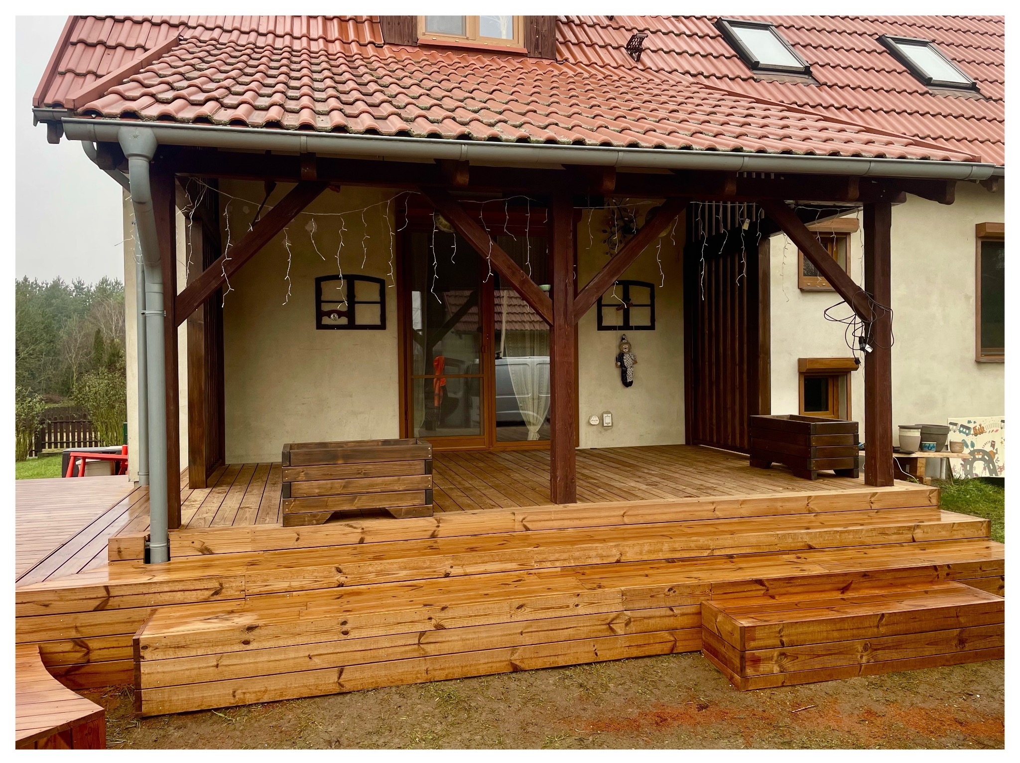 Zadaszony taras z drewna, z niskimi schodami i drewnianymi donicami. Widoczne elementy konstrukcyjne z drewna, oświetlenie dekoracyjne i fragment dachu z dachówką.