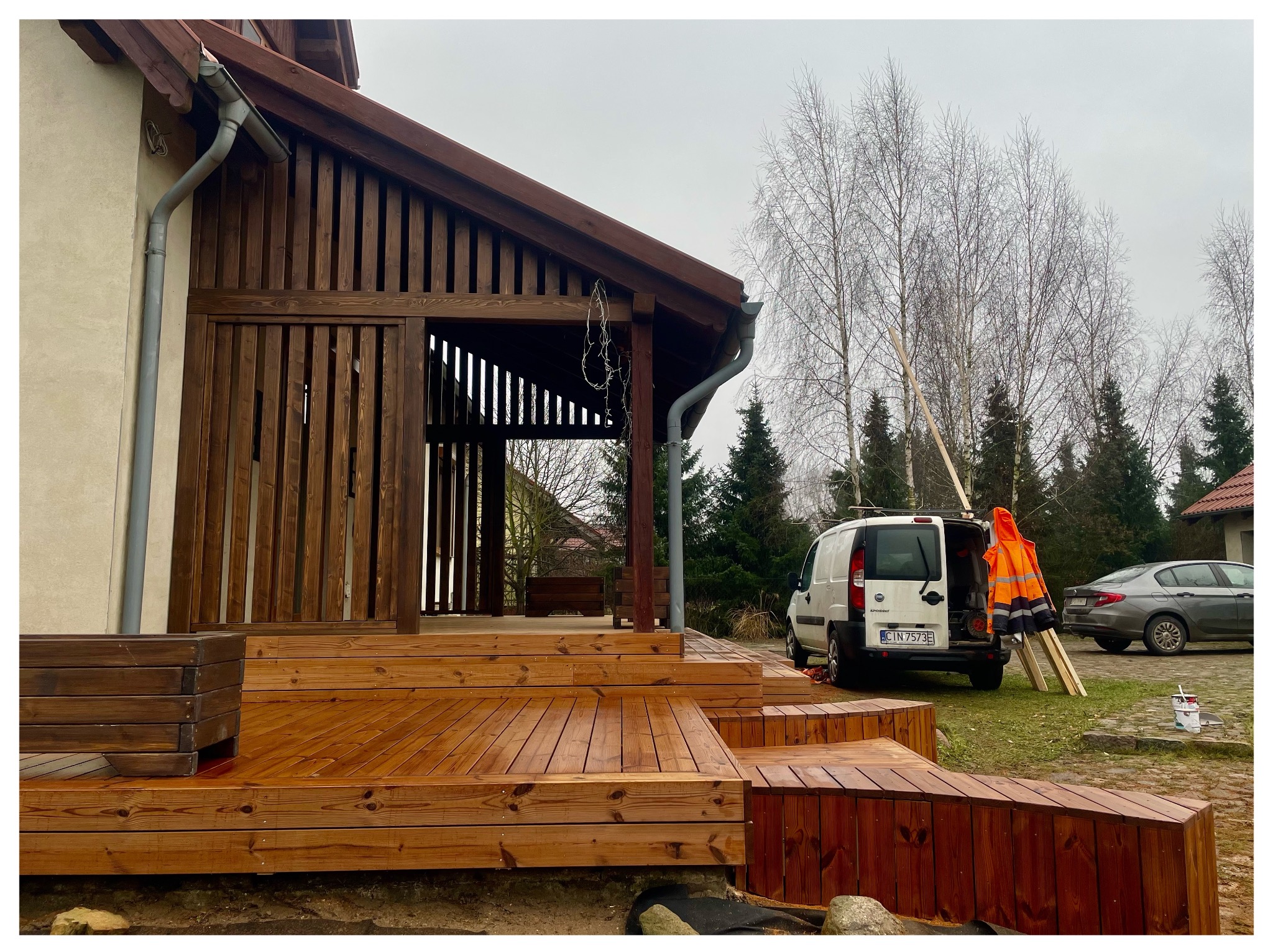 Zadaszony taras z drewna, z widocznymi schodami i elementami konstrukcyjnymi. W tle samochód dostawczy i drzewa bez liści. Widoczne narzędzia i farby sugerują prace wykończeniowe.