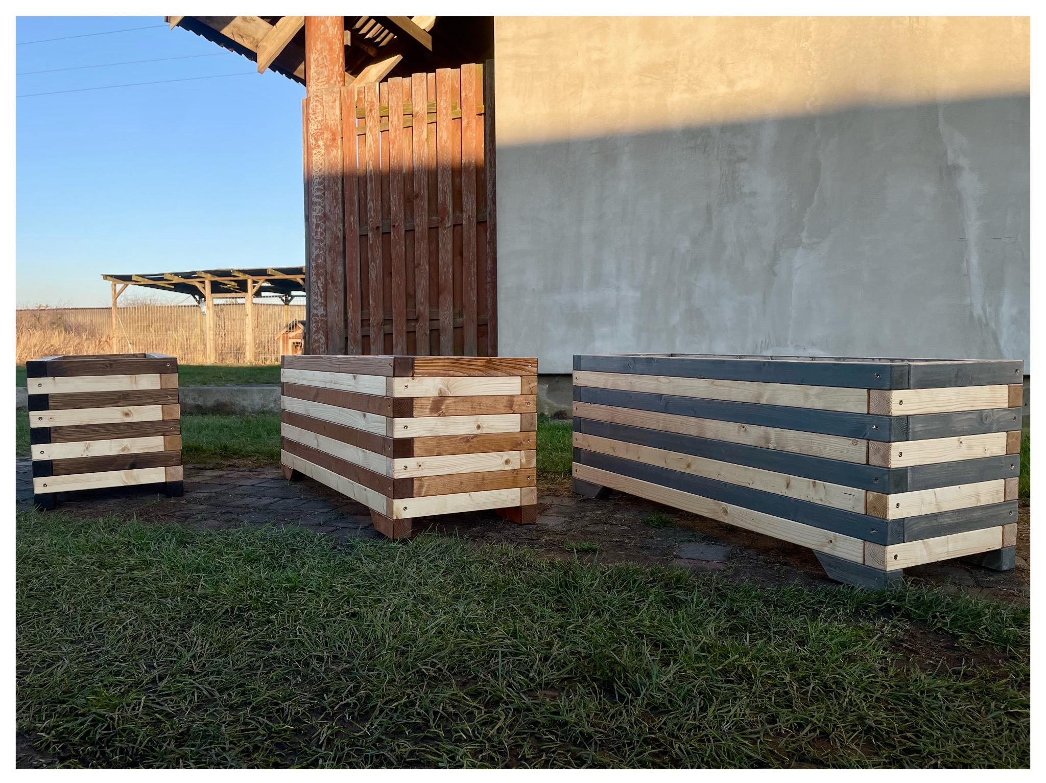 Trzy drewniane skrzynie na rośliny, każda z poziomych desek w innym kolorze, ustawione na trawie przed budynkiem w słoneczny dzień. Widoczne detale łączeń i faktura drewna.