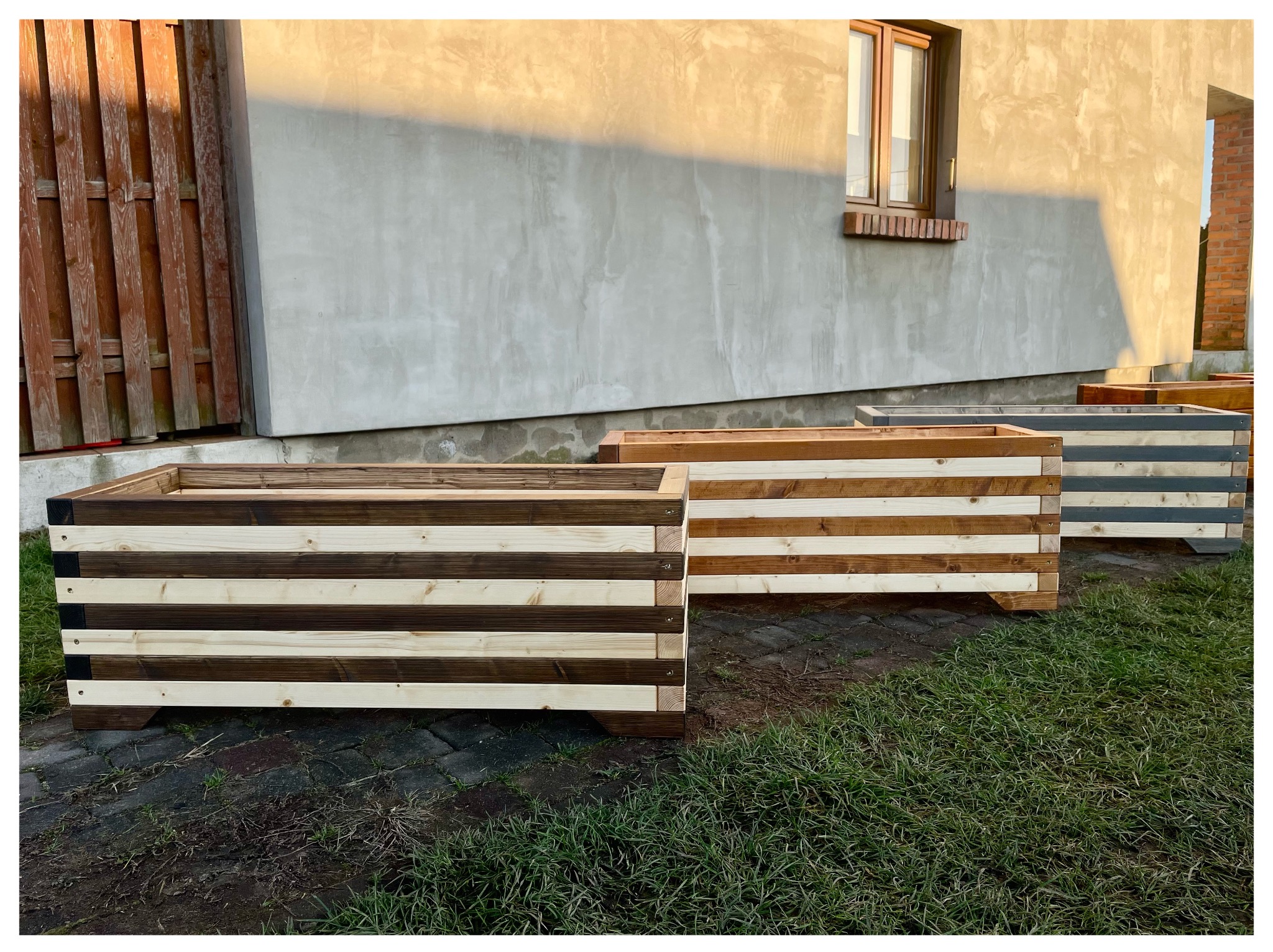 Ręcznie robione, drewniane skrzynie na rośliny w odcieniach brązu i beżu, ustawione na trawie przed budynkiem w słoneczny dzień. Widoczne detale konstrukcji i naturalne usłojenie drewna.