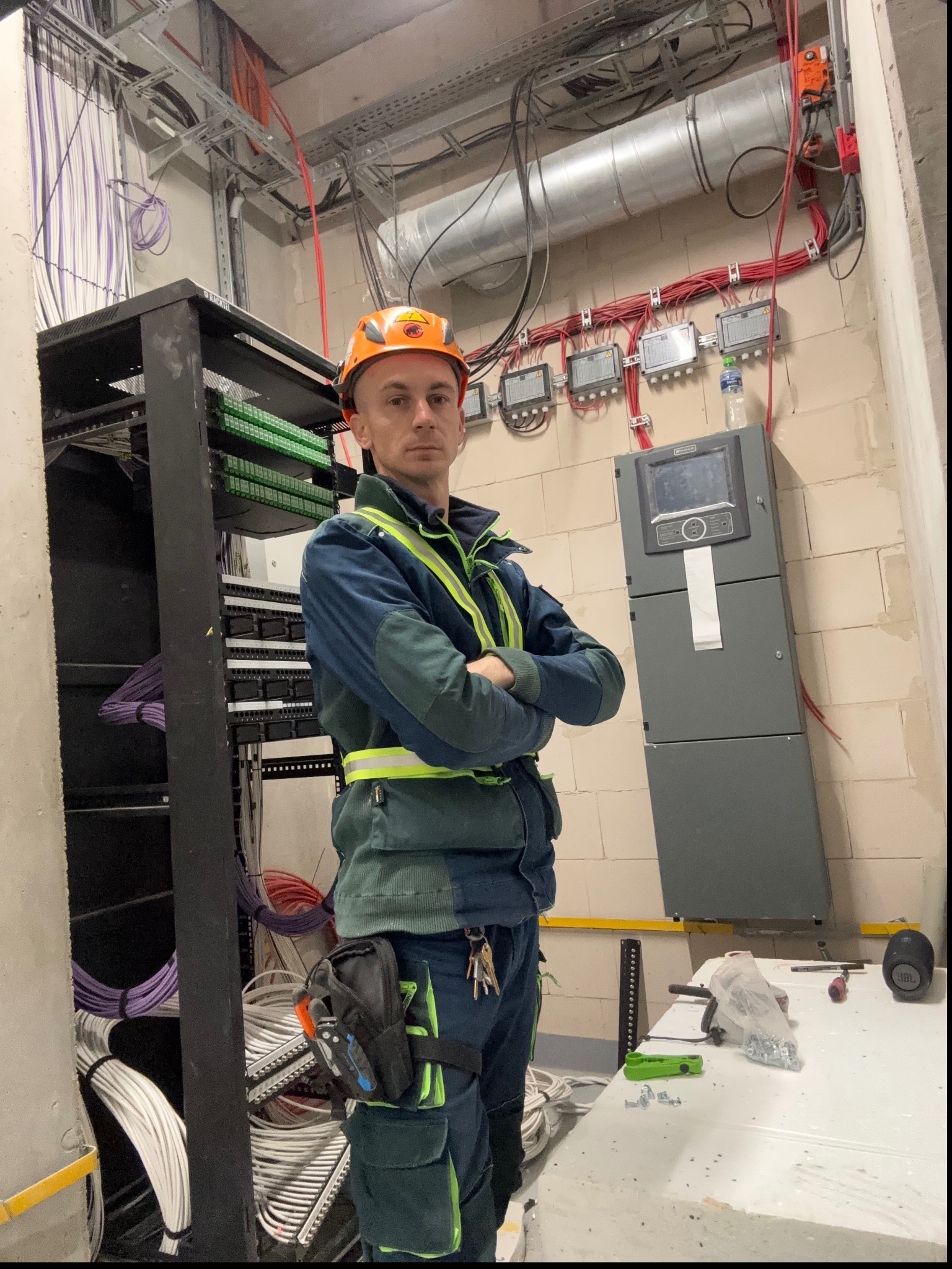 Elektryk w kasku i odblaskowej kamizelce na tle szafy rack z kablami i panelu sterowania, widoczne narzędzia na stole