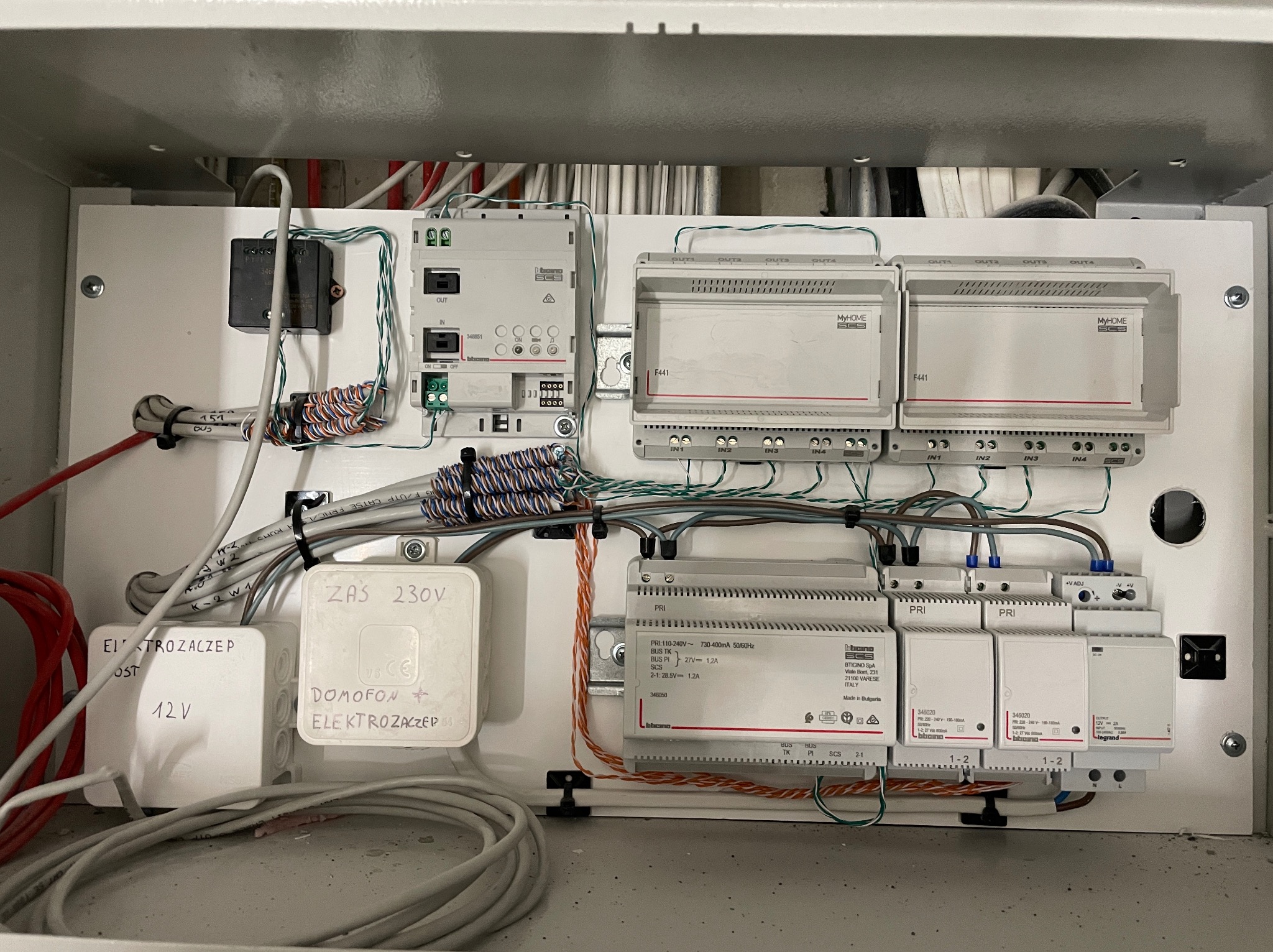 Szafa sterownicza z elementami instalacji domofonowej i elektrozaczepu, widoczne przewody, moduły sterujące i zasilacze, opisane elementy na obudowach.