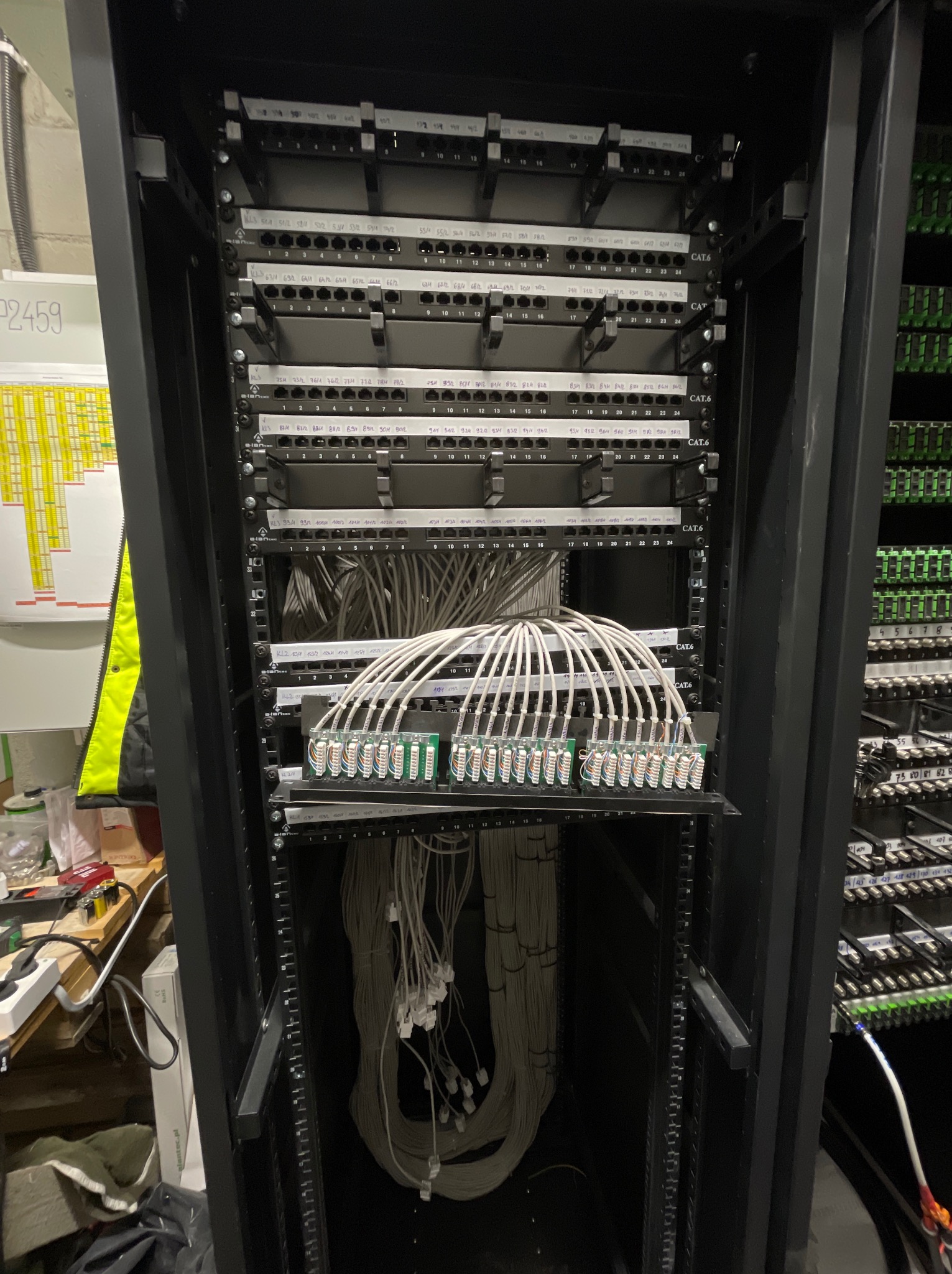 Szafa rack z patch panelem i uporządkowanymi kablami sieciowymi CAT6, widoczne oznaczenia portów i numeracja, z boku widoczne kable i osprzęt elektryczny.