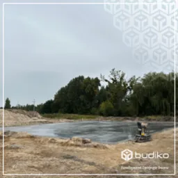 Wylany fundament z betonu wyrównywany zacieraczką mechaniczną na placu budowy, w tle drzewa i pochmurne niebo.