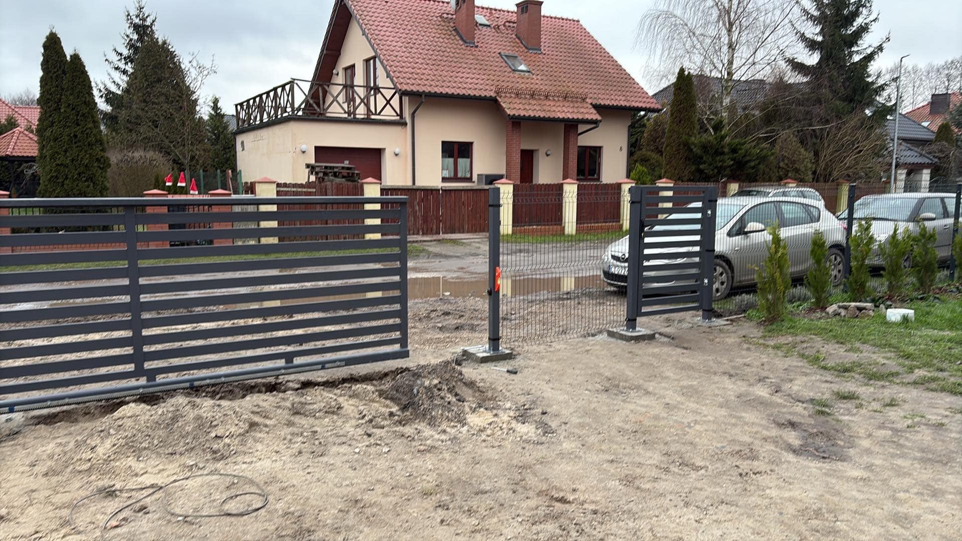 Nowoczesna, pozioma brama wjazdowa z automatyką, w trakcie montażu na posesji z domem jednorodzinnym, widoczne elementy konstrukcyjne i przewody.