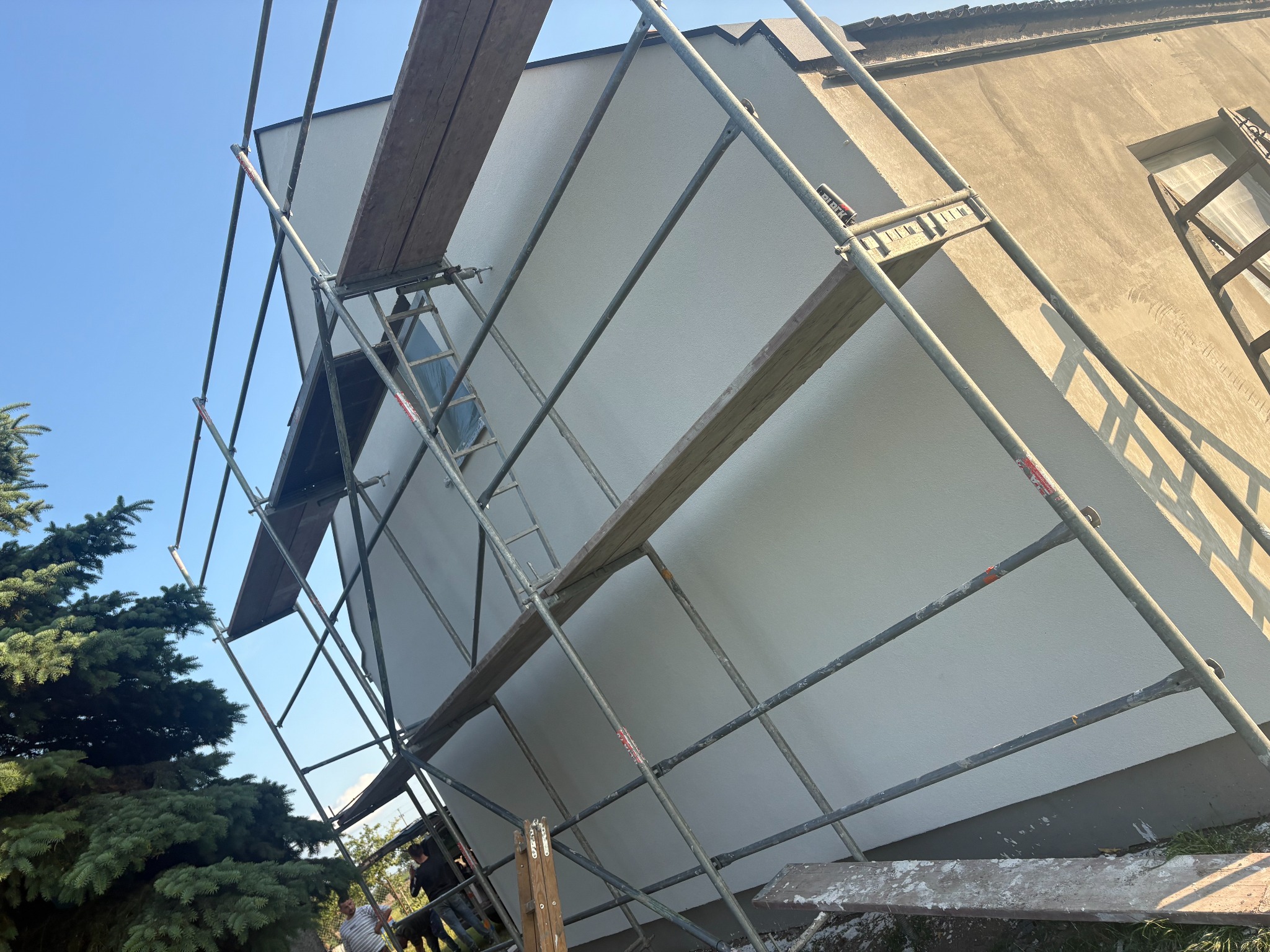 Ocieplona, pomalowana na biało ściana budynku z rusztowaniem. Część ściany w stanie surowym. Widoczne drzewo iglaste i fragment nieba.