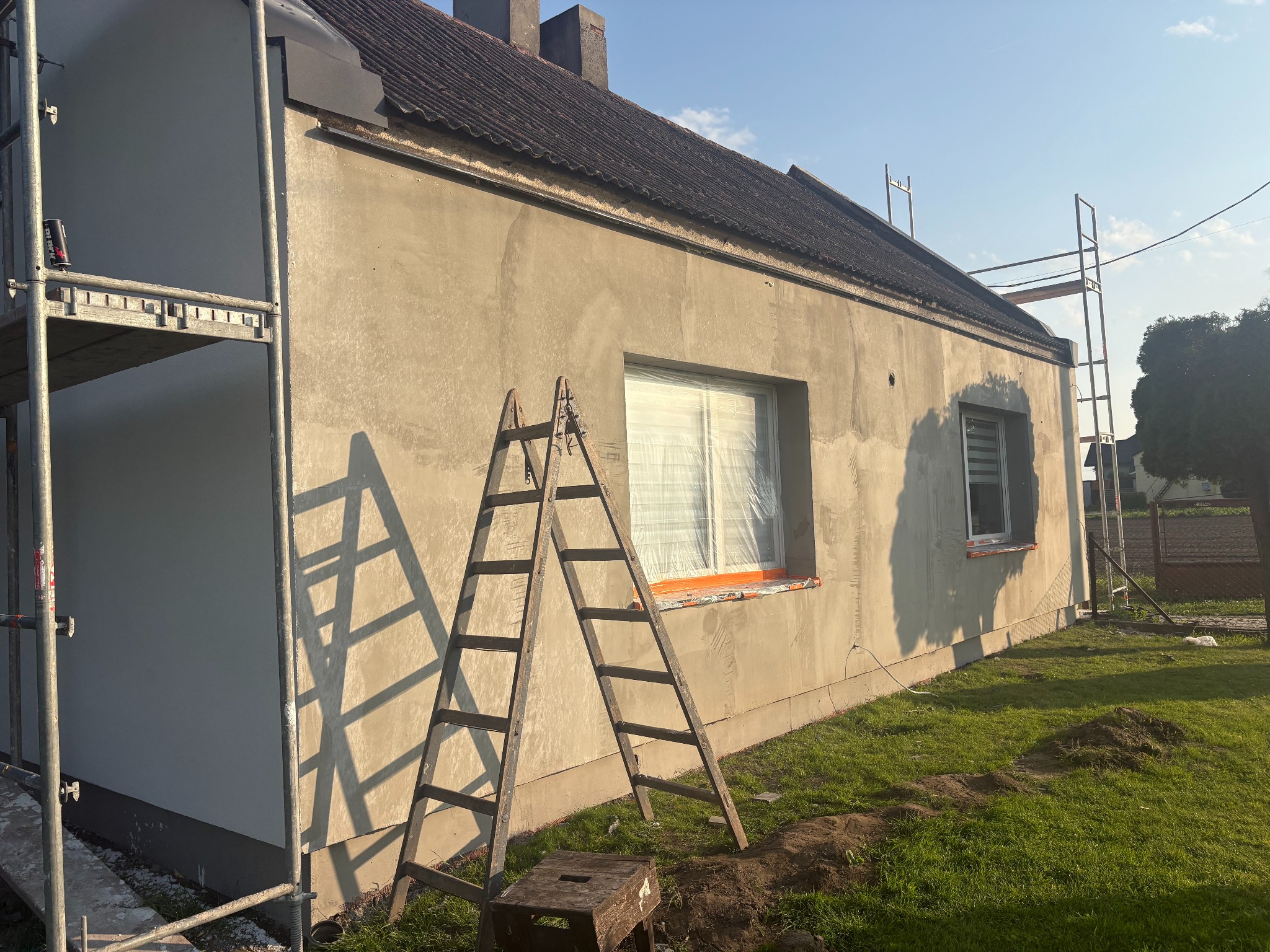 Dom w trakcie ocieplania: część elewacji pomalowana na biało, reszta w szarym tynku. Stoją drabiny i rusztowanie. Okna zabezpieczone folią. Widok na budynek z trawnika.