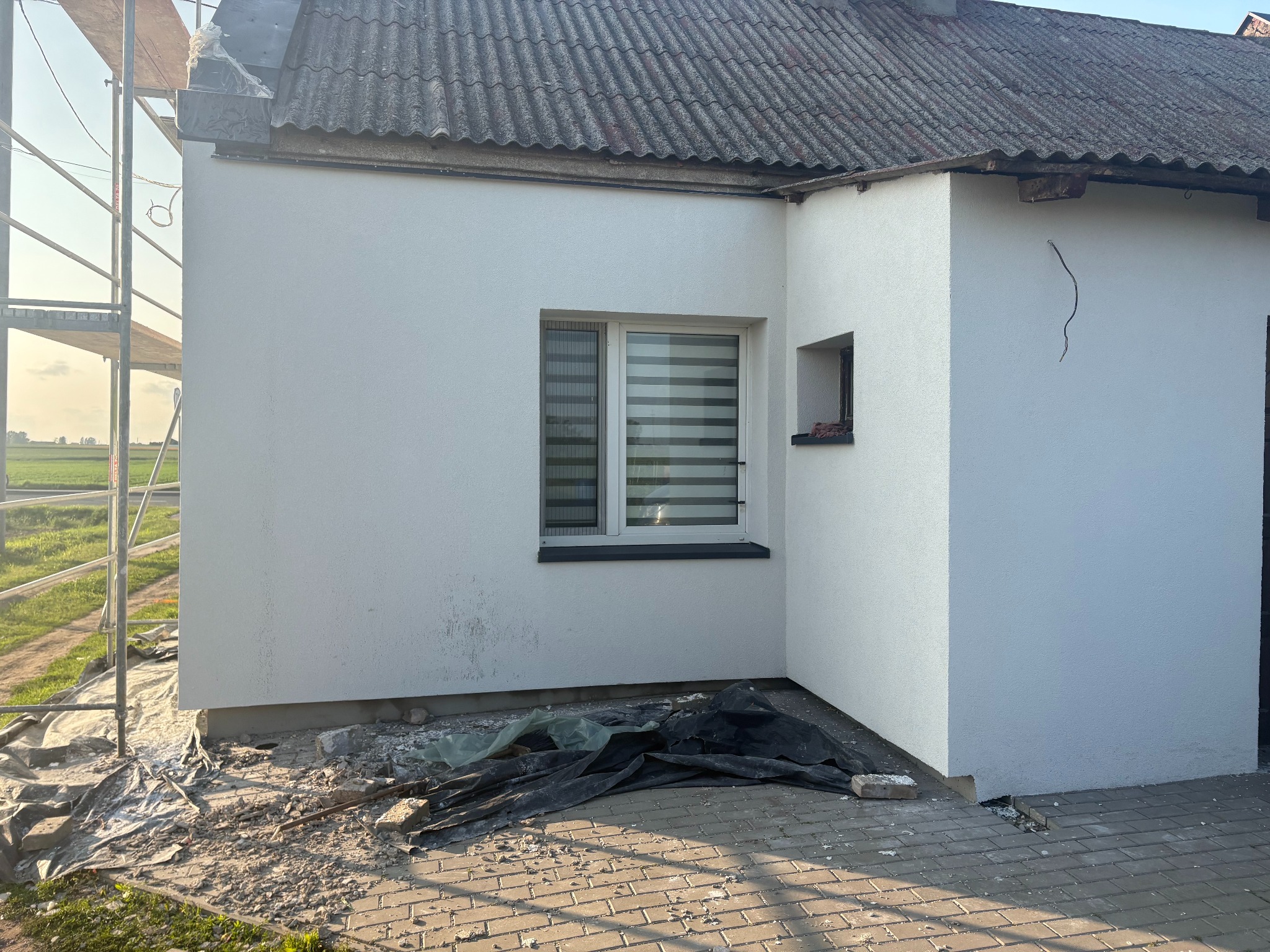 Biała elewacja budynku z szarą dachówką, widoczne rusztowanie i okno z żaluzjami. Na ziemi gruz i folia budowlana. Ujęcie z boku, widać fragment pola w tle.