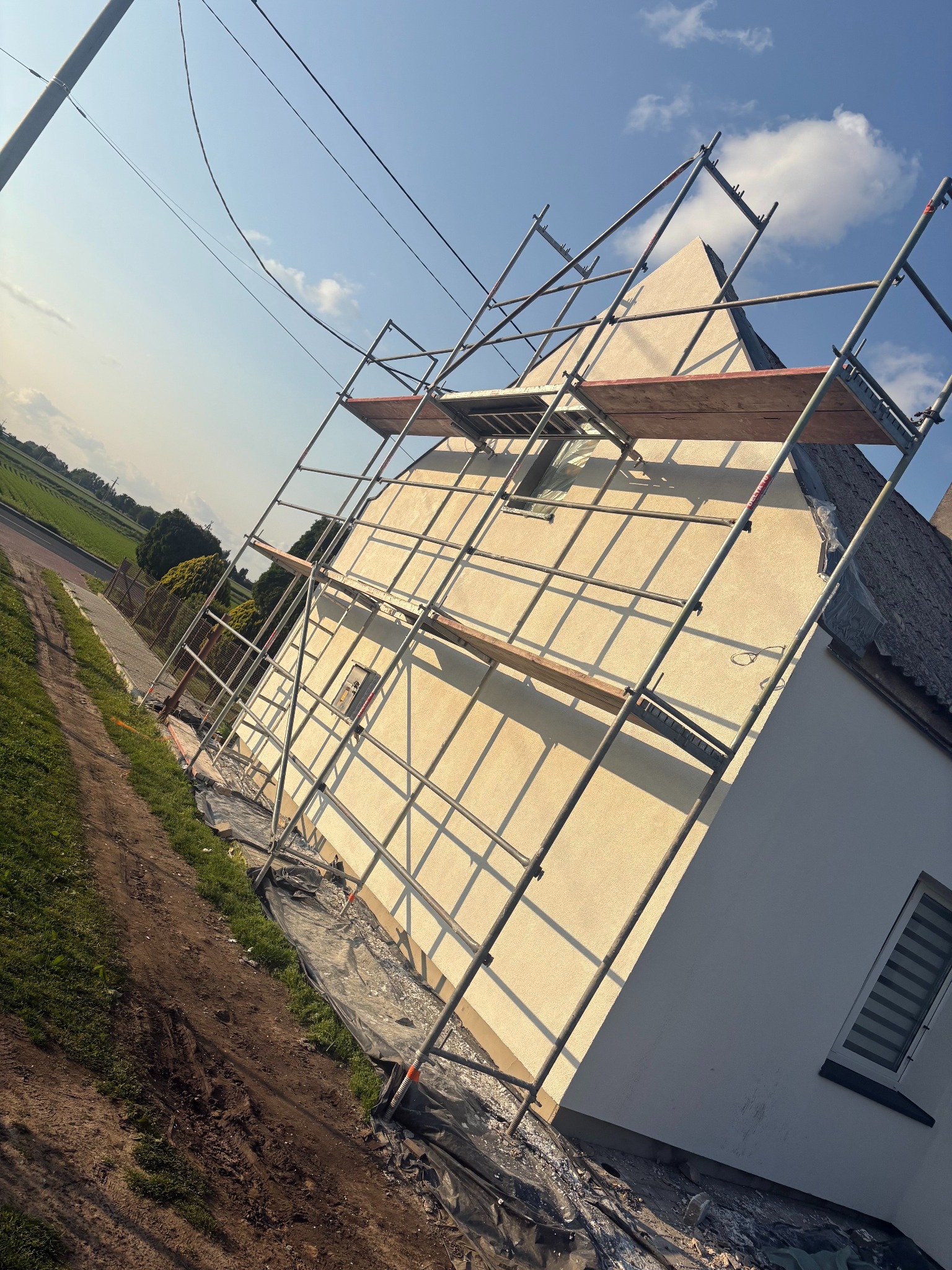 Dom jednorodzinny w trakcie prac elewacyjnych, otoczony rusztowaniem. Widoczne świeżo otynkowane ściany i zabezpieczona folią przestrzeń wokół budynku. Ujęcie z boku budynku.
