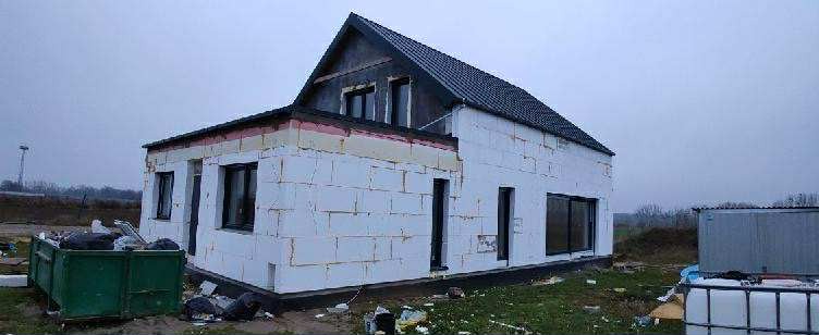 Dom w trakcie ocieplania elewacji styropianem, widoczne okna z czarnymi ramami, dach pokryty dachówką. Na pierwszym planie kontener na odpady i zbiornik na wodę.