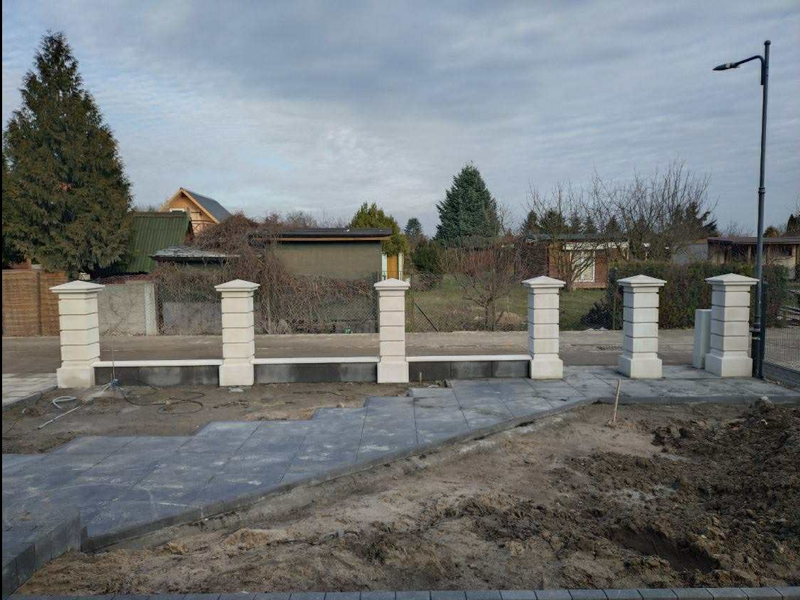Wznoszenie ogrodzenia z betonowych słupków i podmurówki. Częściowo ułożona kostka brukowa na podjeździe. Widoczne elementy instalacji elektrycznej.