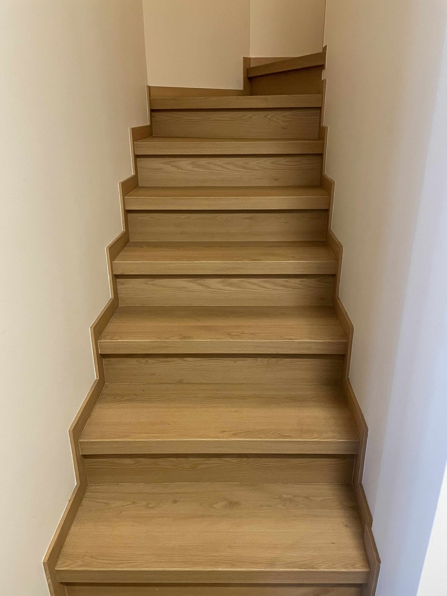 Drewniane schody w jasnym odcieniu dębu, prowadzące w górę, z widoczną strukturą drewna i minimalistycznym wykończeniem przy białej ścianie.