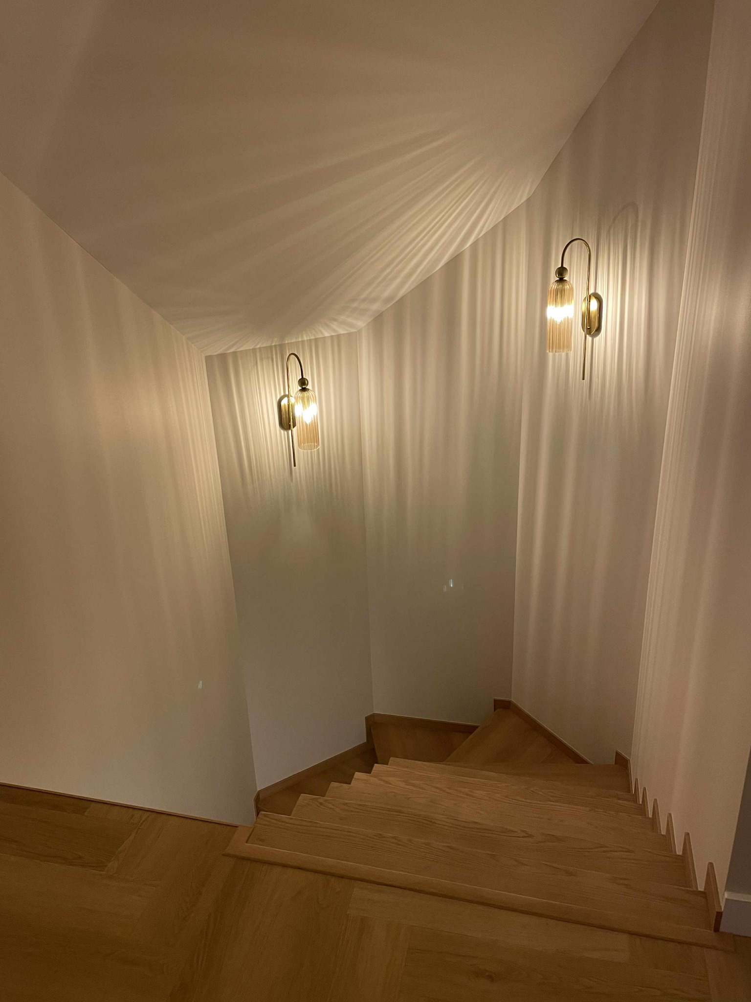 Drewniane schody z oświetleniem ściennym w ciepłych barwach, prowadzące w dół. Parkiet na podłodze, białe ściany. Widok z góry na dół.