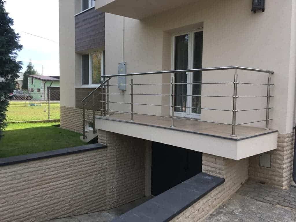 Balkon z metalową balustradą i wejściem do piwnicy w budynku o beżowej elewacji. Widoczne schody, trawnik i fragment budynku sąsiada.