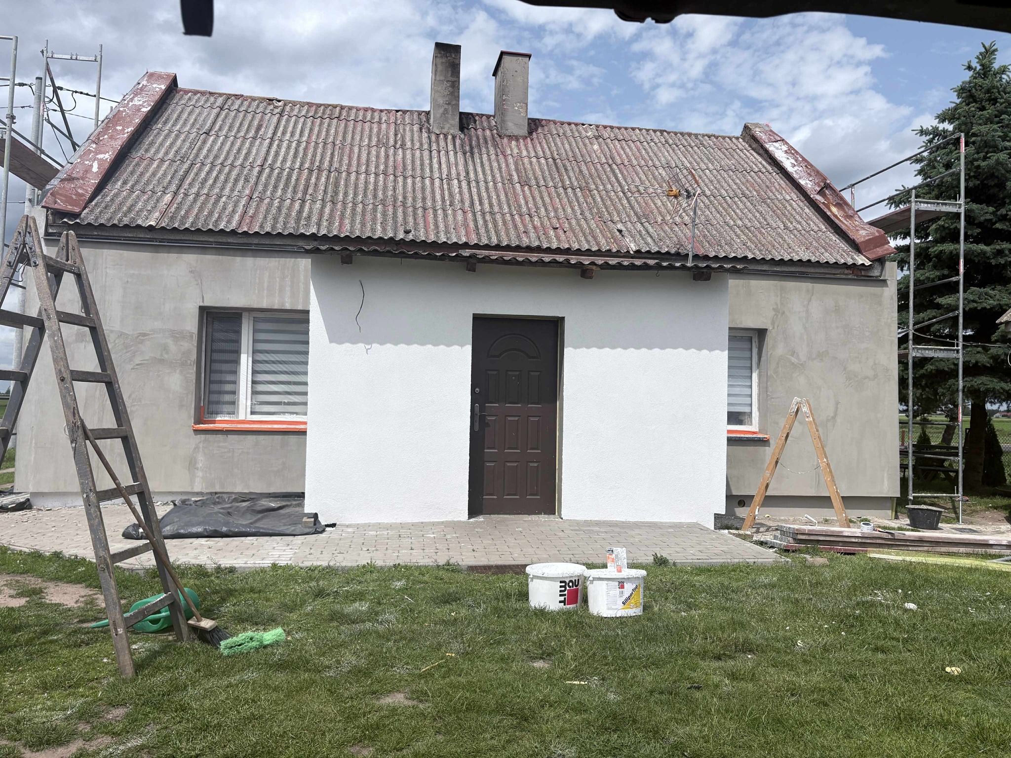 Dom z częściowo otynkowaną elewacją na biało, widoczne rusztowania i drabiny. Część ściany w szarym tynku, reszta biała. Dwa kominy na dachu z szarej dachówki.