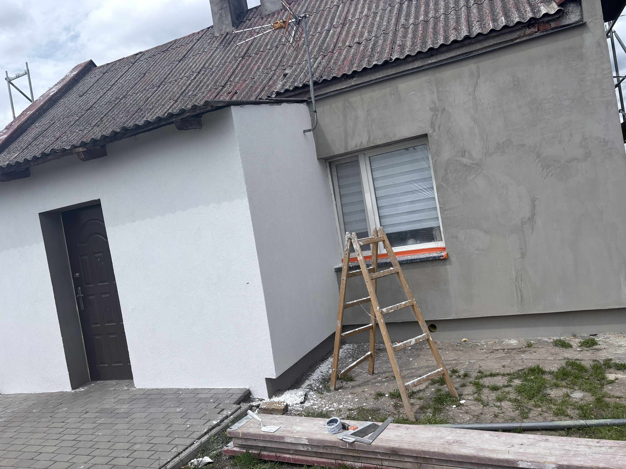 Fragment budynku z ocieploną, pomalowaną na biało częścią elewacji i szarą elewacją w trakcie prac, z drabiną i narzędziami murarskimi w pobliżu.
