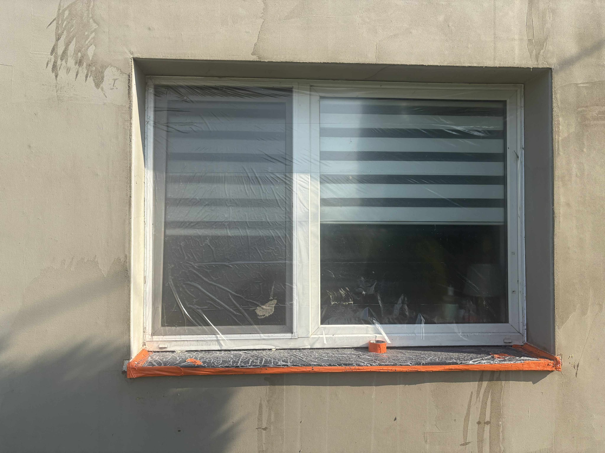 Okna zabezpieczone folią i taśmą malarską podczas nakładania tynku na elewację. Widoczne świeże ślady tynku na ścianie.