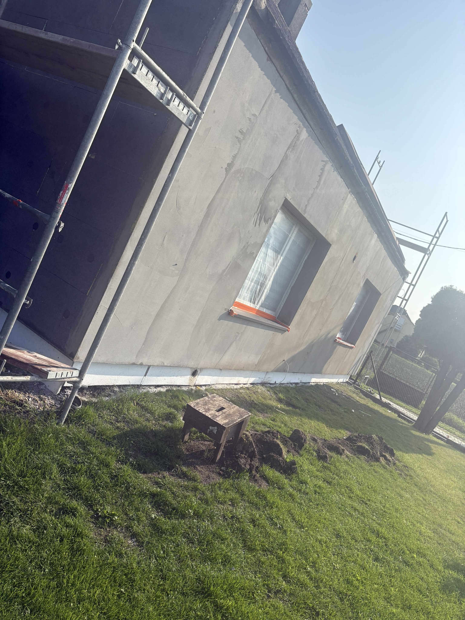 Ocieplona ściana domu w trakcie prac, widoczne rusztowania i zabezpieczone okna. Na trawie leży drewniany taboret.