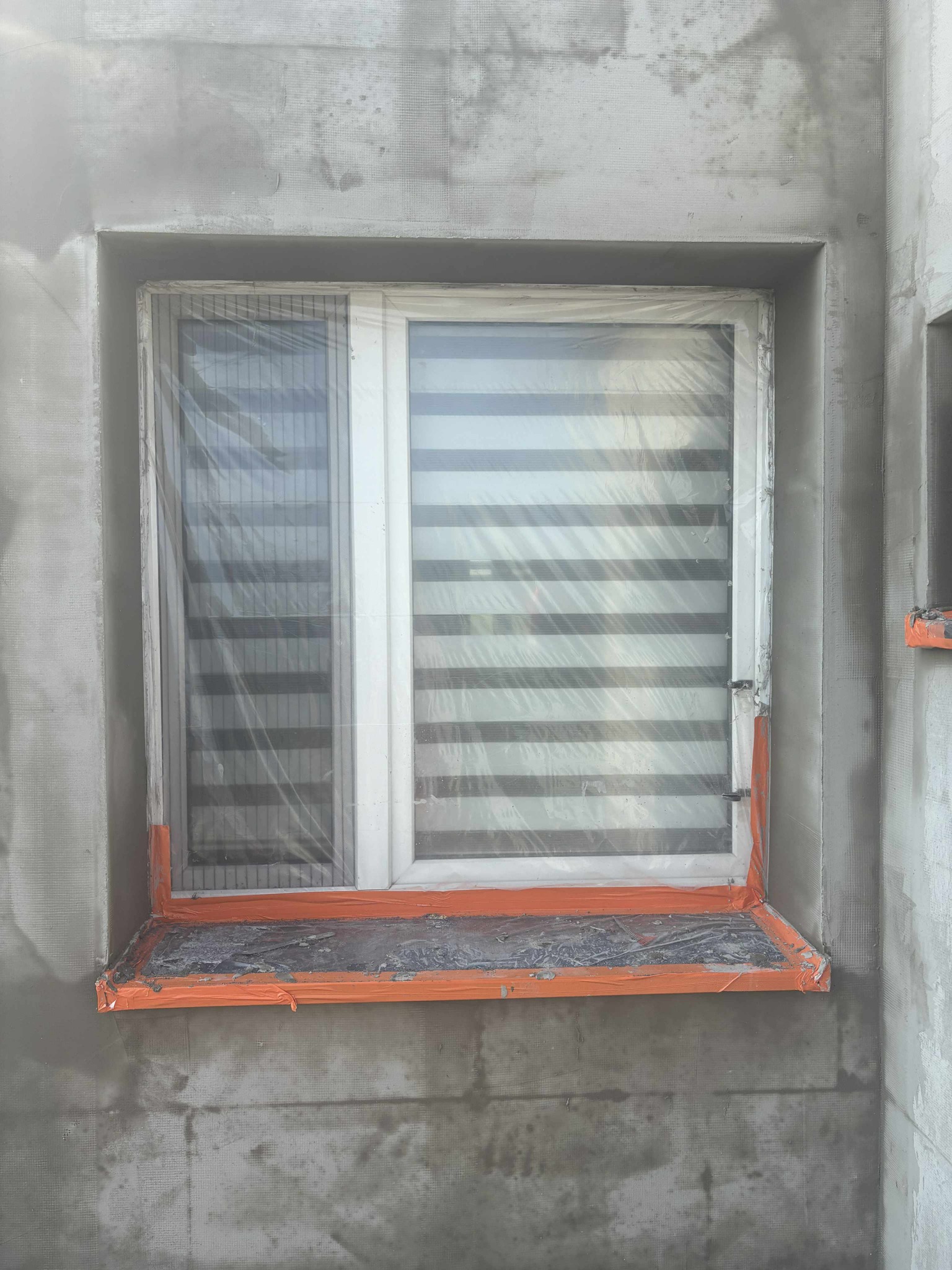 Okna zabezpieczone folią i taśmą malarską podczas prac elewacyjnych. Widoczna siatka pod tynk i surowy tynk na ścianie. Fragment drugiego okna po prawej.