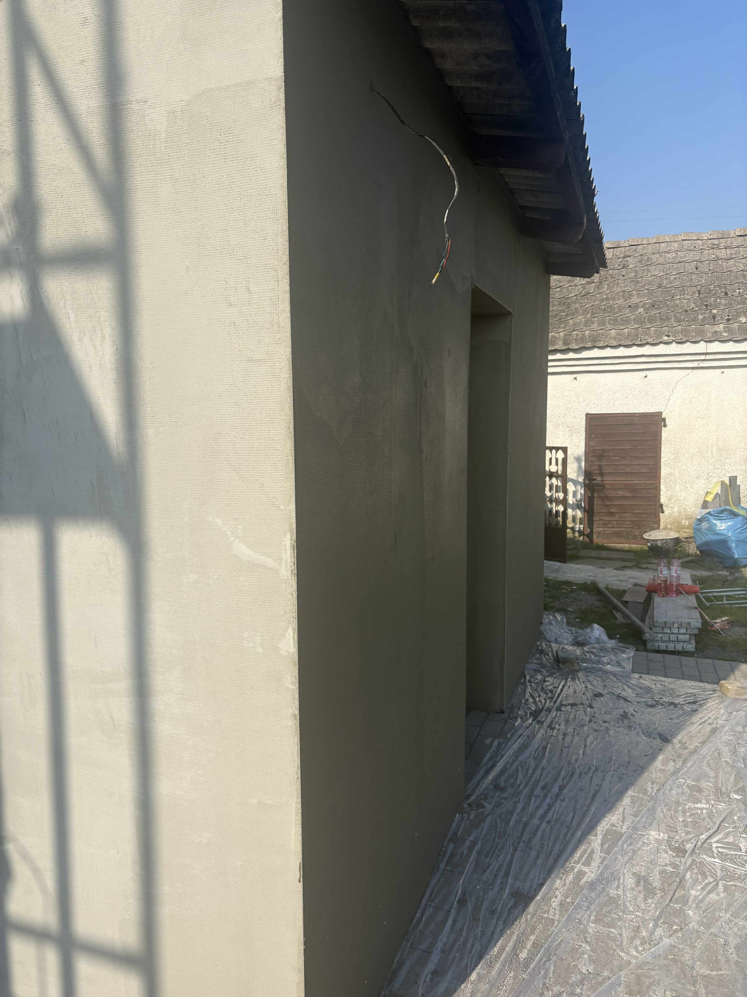 Świeżo otynkowana, beżowa ściana budynku z widocznymi przewodami elektrycznymi, fragmentem dachu i cieniem kratownicy. Prace wykończeniowe w toku.