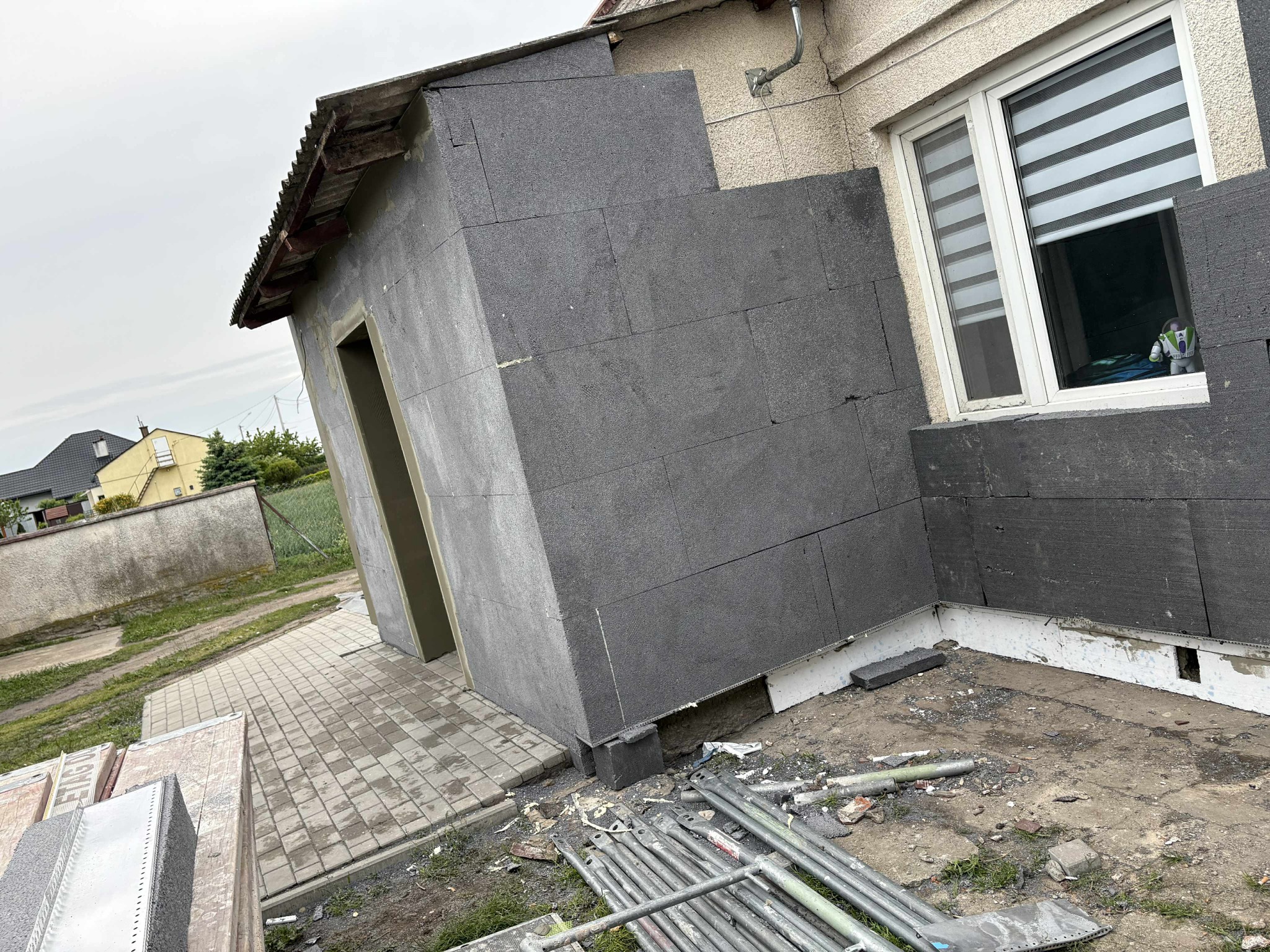 Ocieplony fragment elewacji budynku z szarym styropianem grafitowym, widoczne rusztowania i zabawka w oknie. Prace w toku, widok z dołu na elewację.
