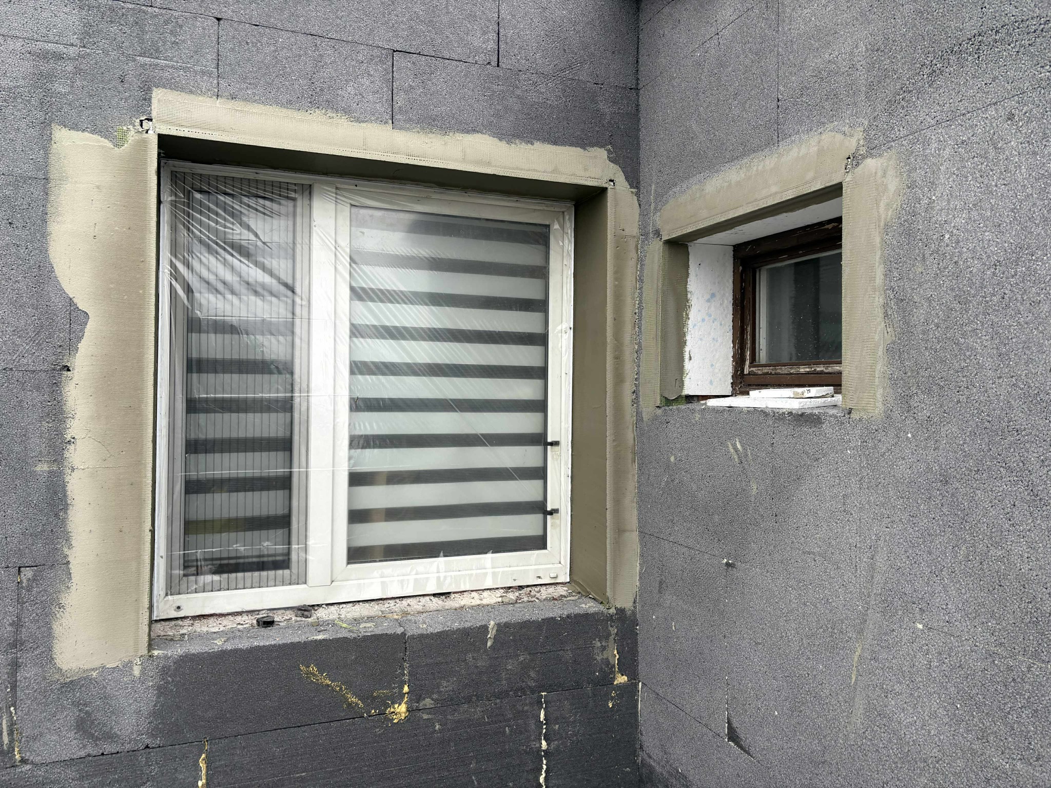 Szare bloczki elewacyjne z oknami w trakcie prac wykończeniowych; jedno okno zabezpieczone folią, drugie starsze. Widoczne detale obróbki okiennej.