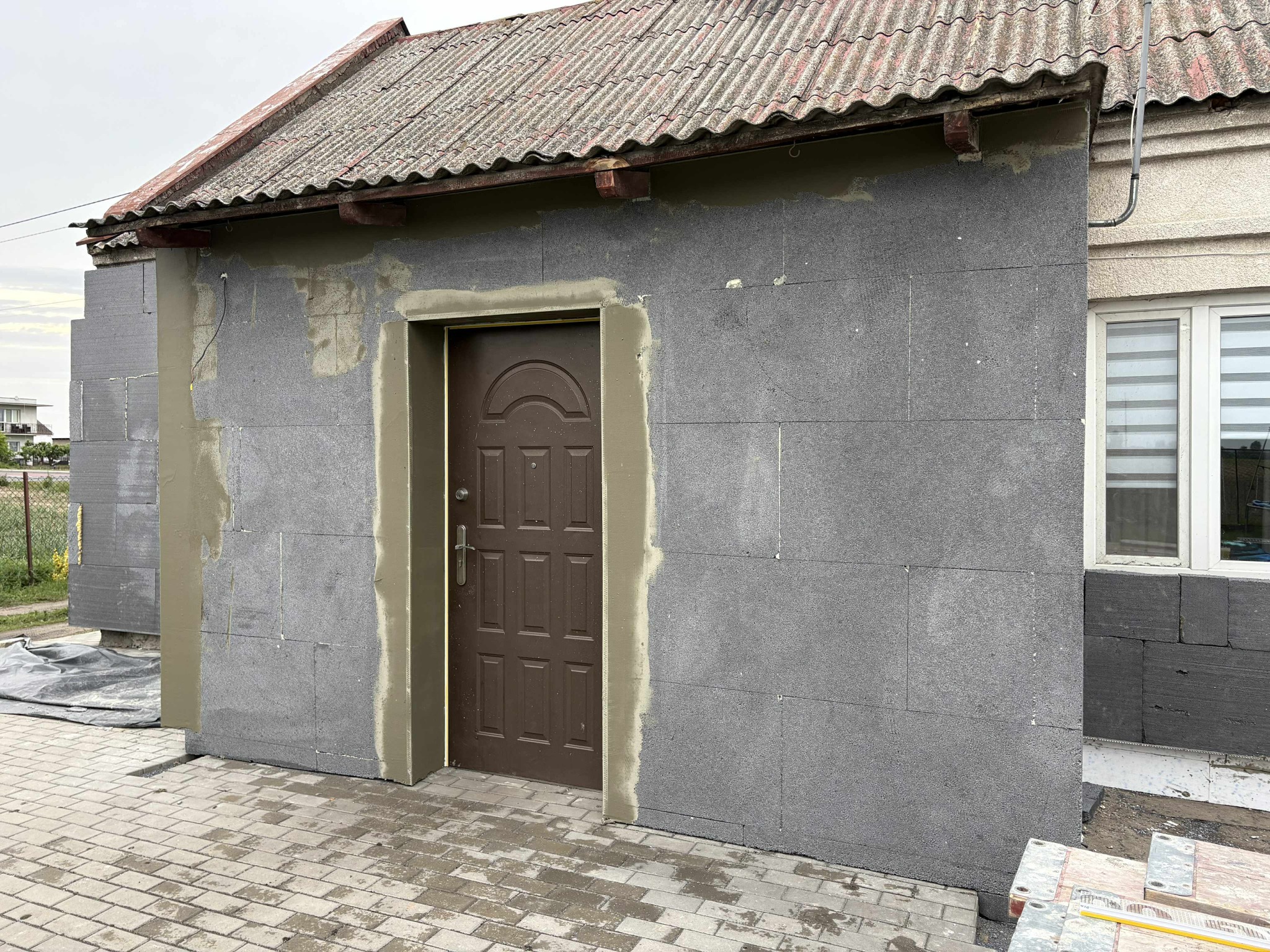 Montaż szarej izolacji termicznej na ścianie budynku z brązowymi drzwiami wejściowymi, widoczne elementy rusztowania i kostka brukowa. Prace ociepleniowe w toku.