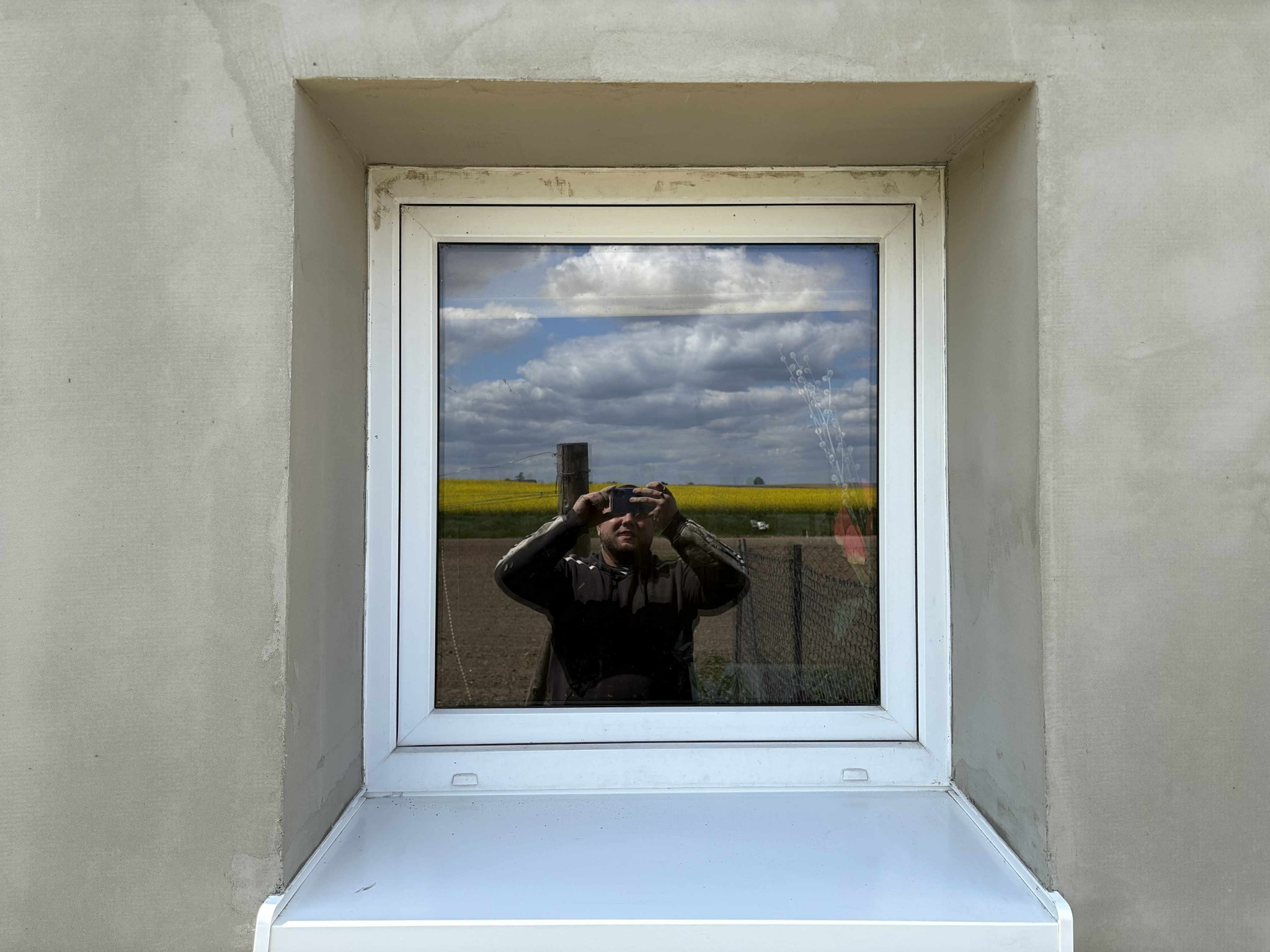 Okna po montażu w surowym otoczeniu, ściana po szpachlowaniu, widoczne niedoskonałości i odbicie fotografa w szybie. Krajobraz w tle za oknem.