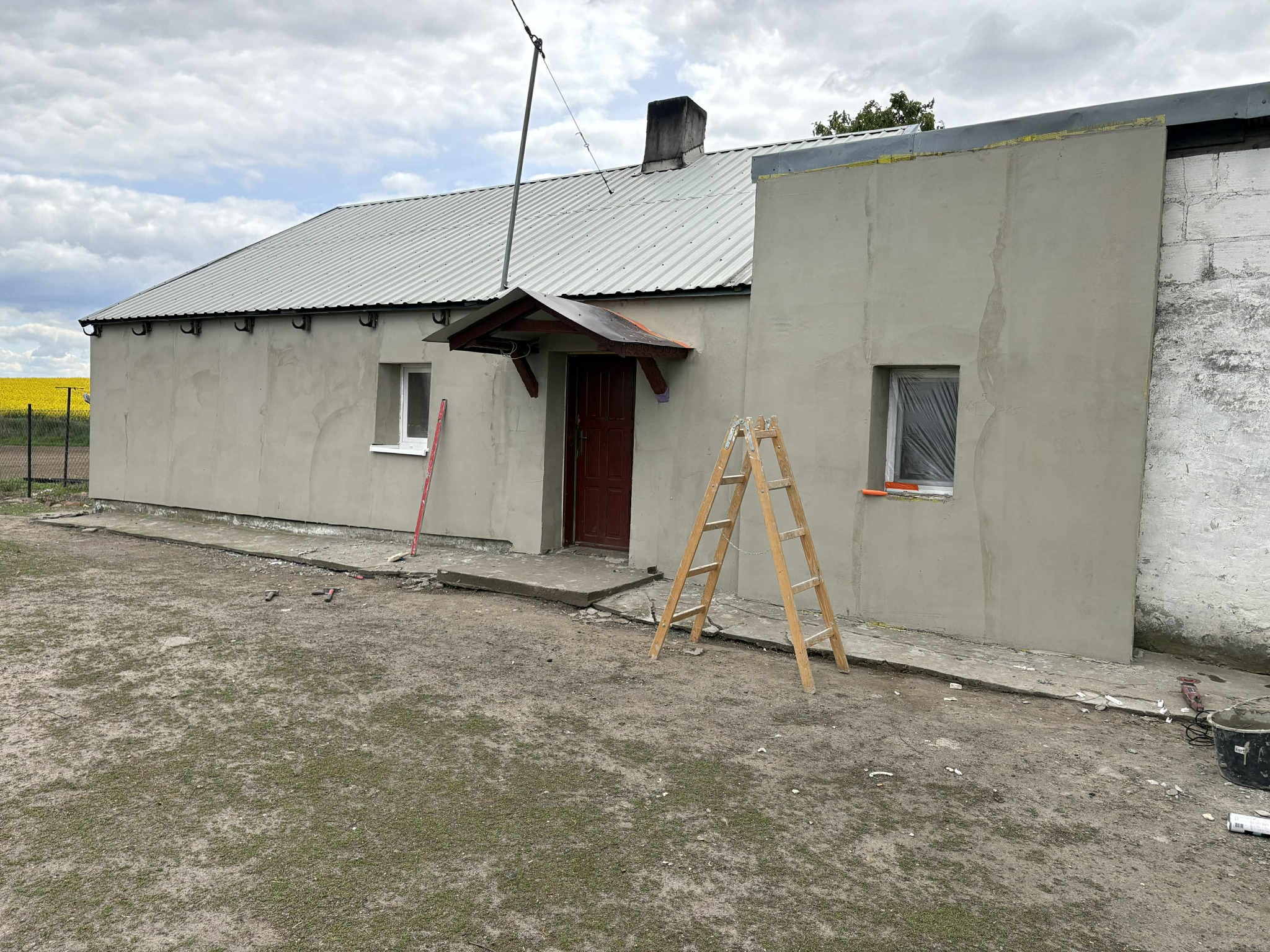 Budynek po nałożeniu szarego tynku, z drewnianą drabiną opartą o ścianę. Widoczne okna zabezpieczone folią, fragment dachu i otoczenie budynku. Prace wykończeniowe w toku.