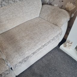 Maya Bee Cleaning Ewa Mendyk - Fragment jasnej kanapy z wzorem w kratkę na obiciu, stojącej na szarym dywanie, z tapetą w kwiaty w tle oraz dekoracyjnym elementem na białej półce.