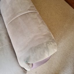 Maya Bee Cleaning Ewa Mendyk - Fragment jasnej, tapicerowanej kanapy z widocznym zaciekłym plamą, częściowo wyczyszczona, na tle beżowego dywanu i fragmentu drewnianego mebla.