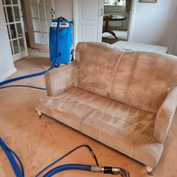 Maya Bee Cleaning Ewa Mendyk - Beżowa kanapa z widocznymi śladami czyszczenia, niebieski odkurzacz piorący z grubymi wężami na beżowej wykładzinie, w tle drzwi z szybkami i obraz na ścianie.