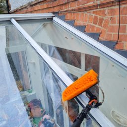 Maya Bee Cleaning Ewa Mendyk - Mycie przeszklonego dachu oranżerii za pomocą szczotki na teleskopowym wysięgniku, widoczne krople wody i refleksy na szybach, w tle ceglana ściana budynku.