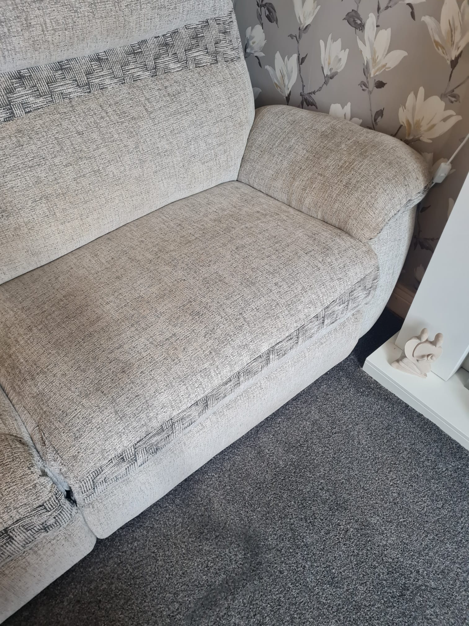 Fragment jasnej kanapy z wzorem w kratkę na obiciu, stojącej na szarym dywanie, z tapetą w kwiaty w tle oraz dekoracyjnym elementem na białej półce.