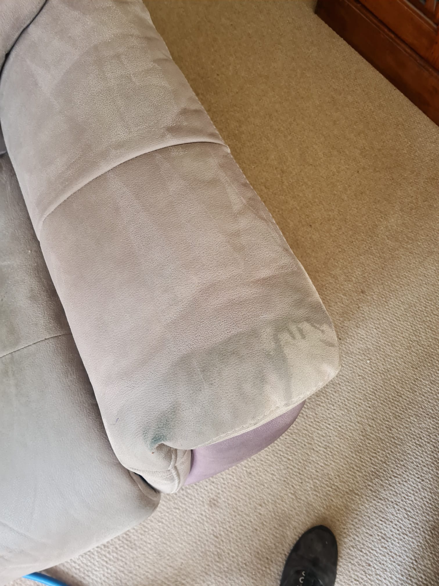 Fragment jasnej, tapicerowanej kanapy z widocznym zaciekłym plamą, częściowo wyczyszczona, na tle beżowego dywanu i fragmentu drewnianego mebla.