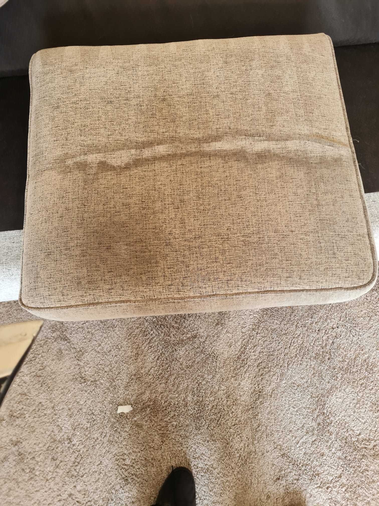 Beżowy, tapicerowany podnóżek z widocznymi plamami na powierzchni, ustawiony na dywanie o podobnej kolorystyce.