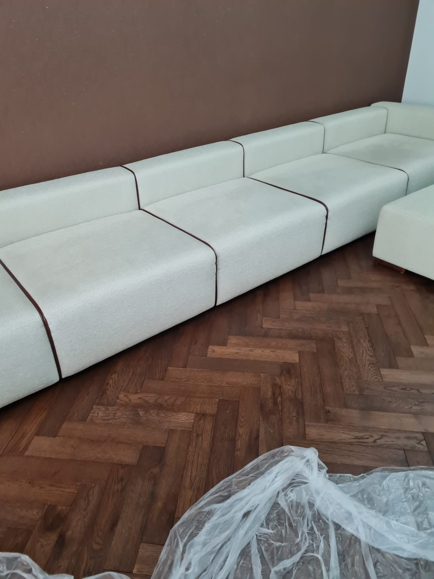 Jasna, modułowa sofa z brązowymi akcentami na parkiecie ułożonym w jodełkę, częściowo przykryta folią ochronną.